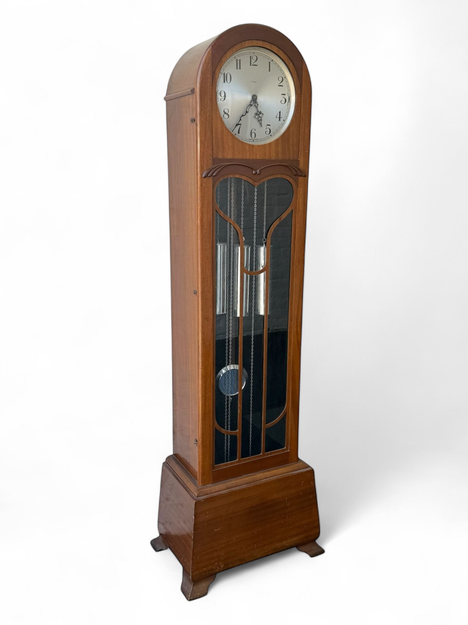 English- Art deco mahogany 8-day longcase clock, with an arched top and fill length fully glazed door beneath, on a short plinth with bracket feet, three train chain driven movement with visible stainless steel cased weights and pendulum, silvered dial with Arabic numerals, minute track and pierced steel hands, quarter chiming movement with  Westminster/ Whittington/ strike/silent selection.

