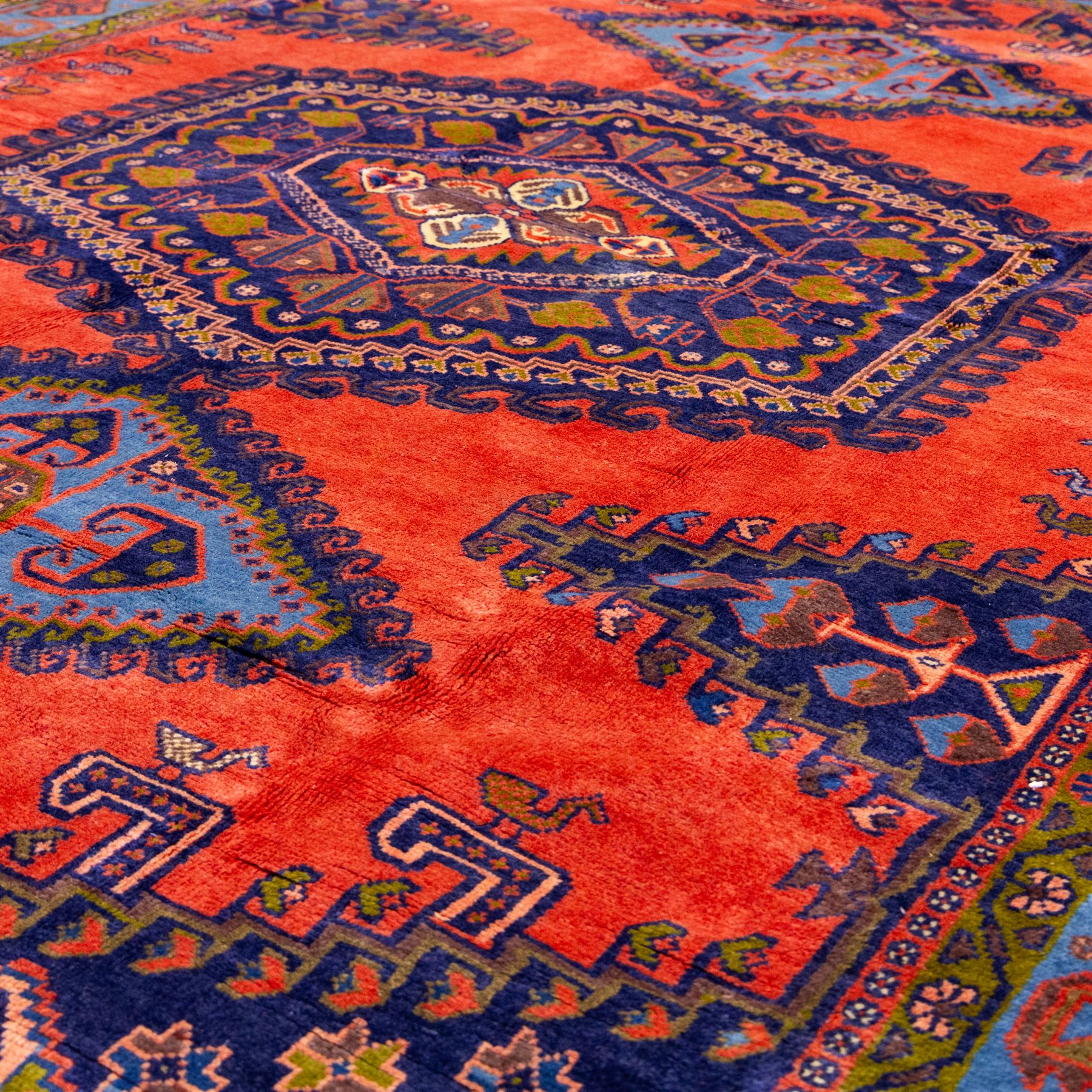 West Persian Veis hand-knotted rug, coral red field decorated with a series of bold geometric lozenge medallions in shades of indigo and sky blue surrounded by angular tribal motifs and stylized animal and plant forms framed by a deep indigo border of geometric ornament and multiple guard stripes, cotton fringe ends 