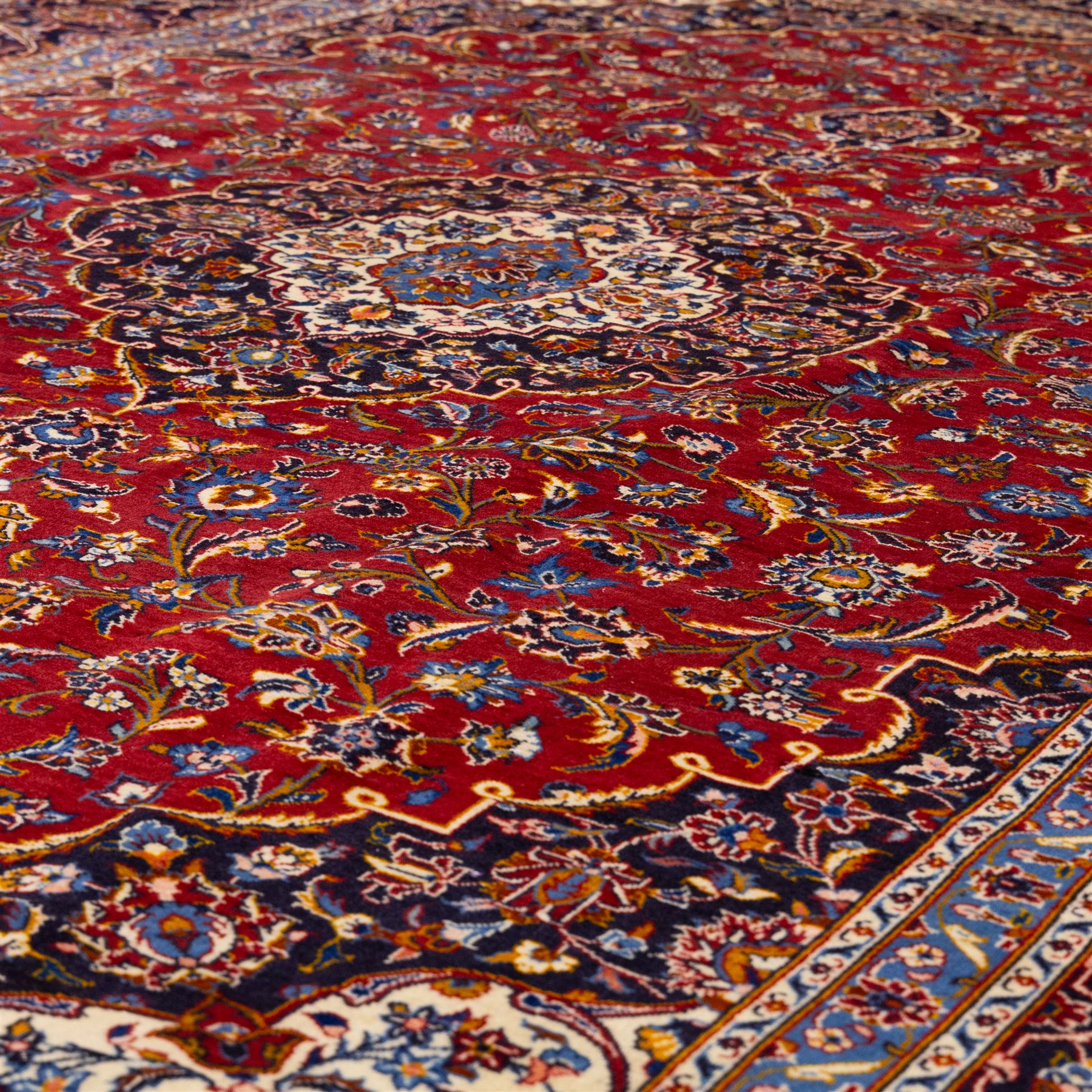 Central Persian Kashan hand-knotted carpet, rich crimson red field with a central cusped medallion with finely drawn floral sprays surrounded by dense scrolling vine work and palmettes, framed by a broad indigo ground border of flowering arabesques flanked by multiple guard stripes, in a palette of red, blue, ivory, sage green, and soft ochre tones, with cotton fringe ends    
