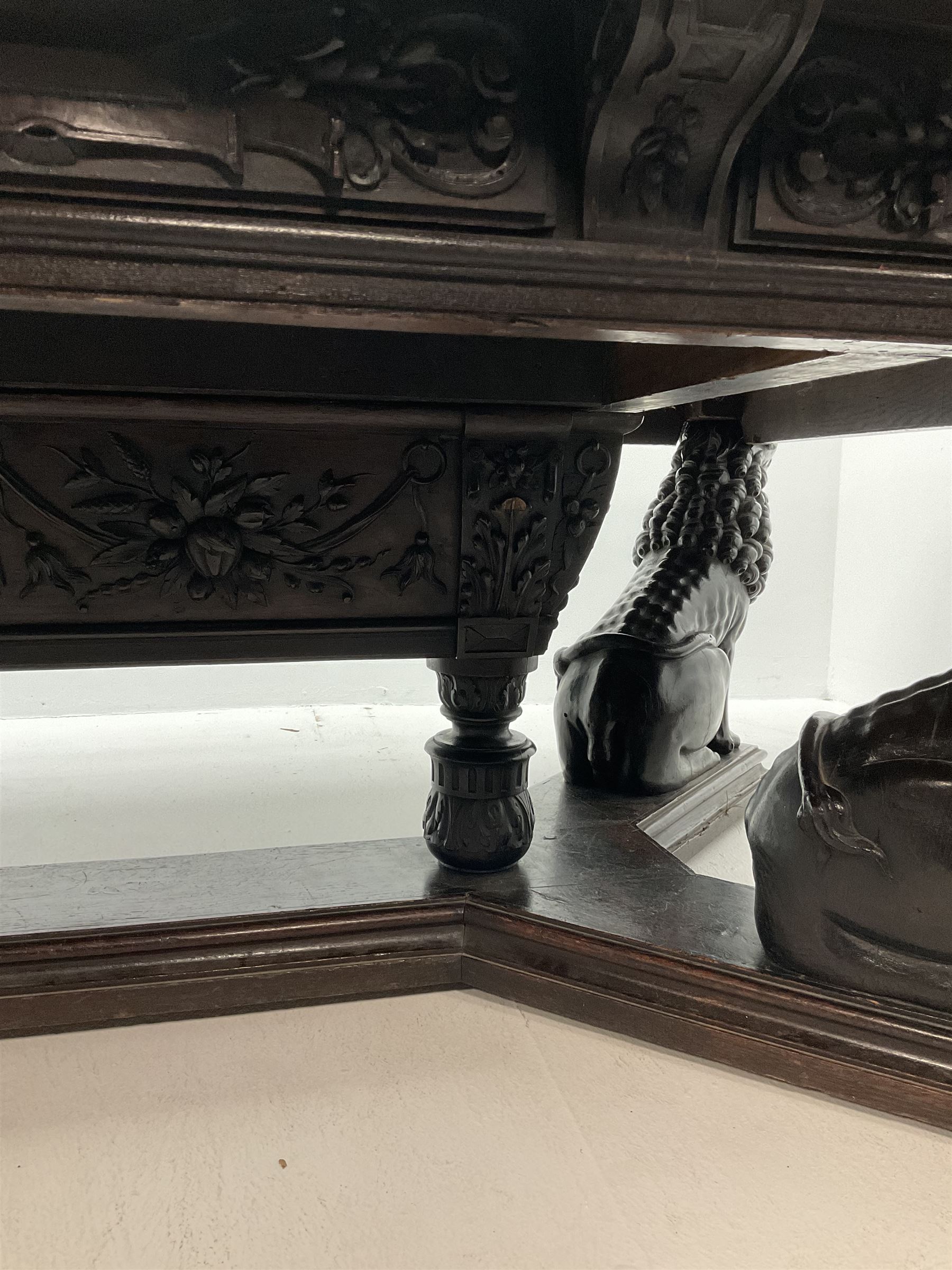 Large 19th century carved oak library table, the rectangular top with lunette carved edge over cartouche panelled and carved  scroll frieze, the central tapering rectangular support relief carved with masks and garlands, on four carved seated lion supports at each corner, on flat angular stretcher with recessed castors stamped ‘Bartholomew & Fletcher, London’