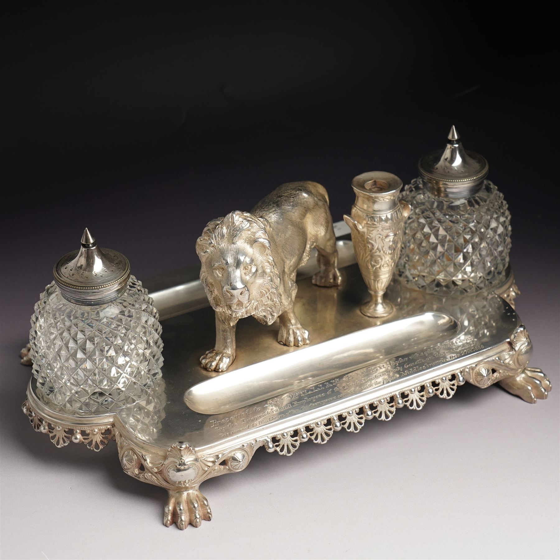 19th century Walker & Hall, silver plated inkstand, oblong and on four paw feet, the border decorated with pierced shell design, each side with a pen-rest, with two cut-glass bottles and central lion and urn finials, engraved to the front with an inscription, H15cm, L30cm