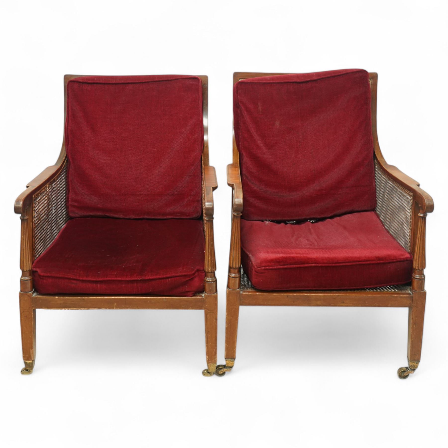 Pair of early 20th-century mahogany Bergère armchairs in the manner of Gillows, the back and seat with woven cane panels with loose red velvet cushions, the frame with swept arms with tapered and reeded uprights, raised on tapered square-section front legs terminating in brass cup castors 