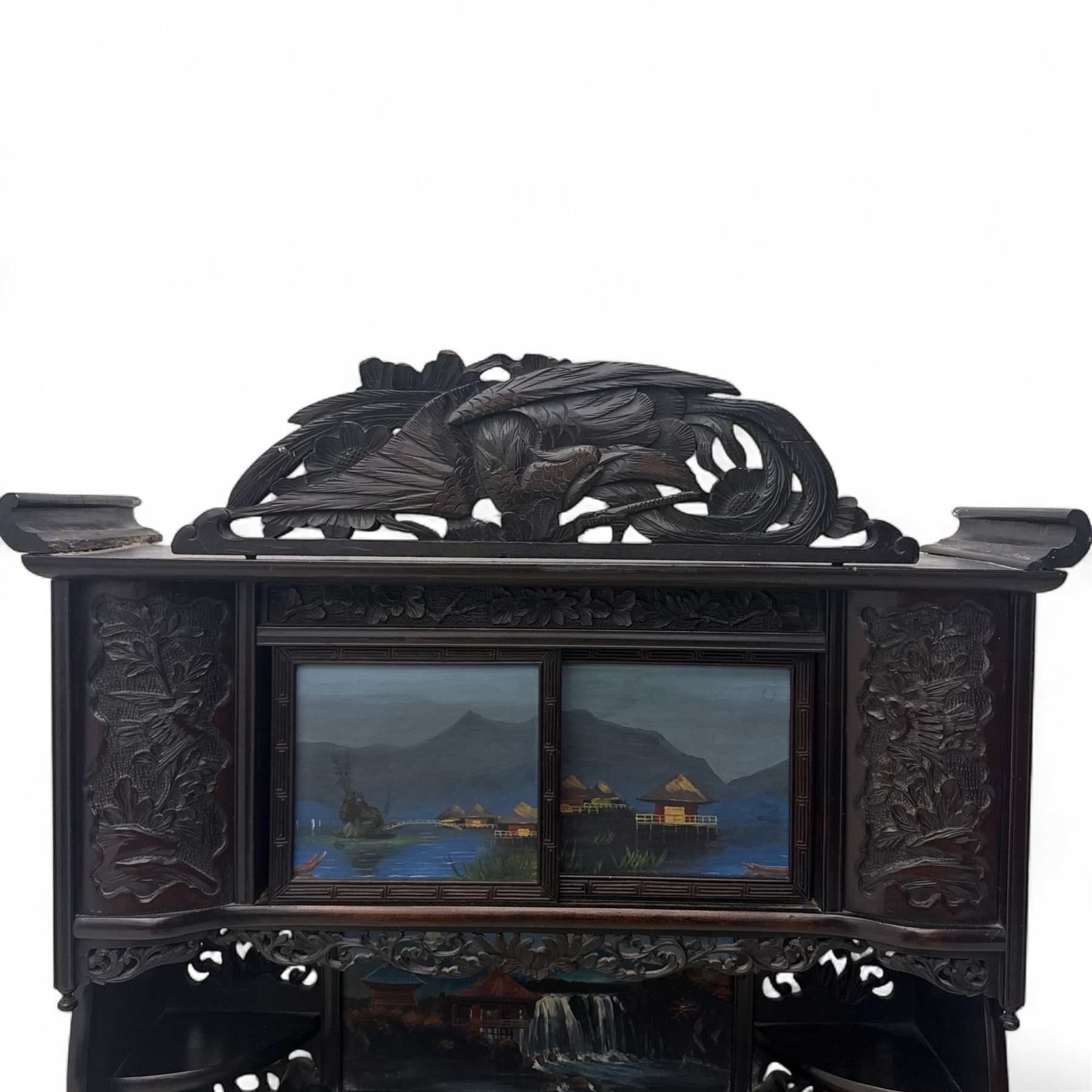 Early 20th century Japanese Meiji style carved hardwood cabinet, of symmetrical tiered form with pierced foliate fretwork panels and phoenix crest, fitted with sliding and hinged doors inset with hand-painted landscape panels depicting Mount Fuji, waterfalls, and lakeside dwellings, raised on shaped bracket supports