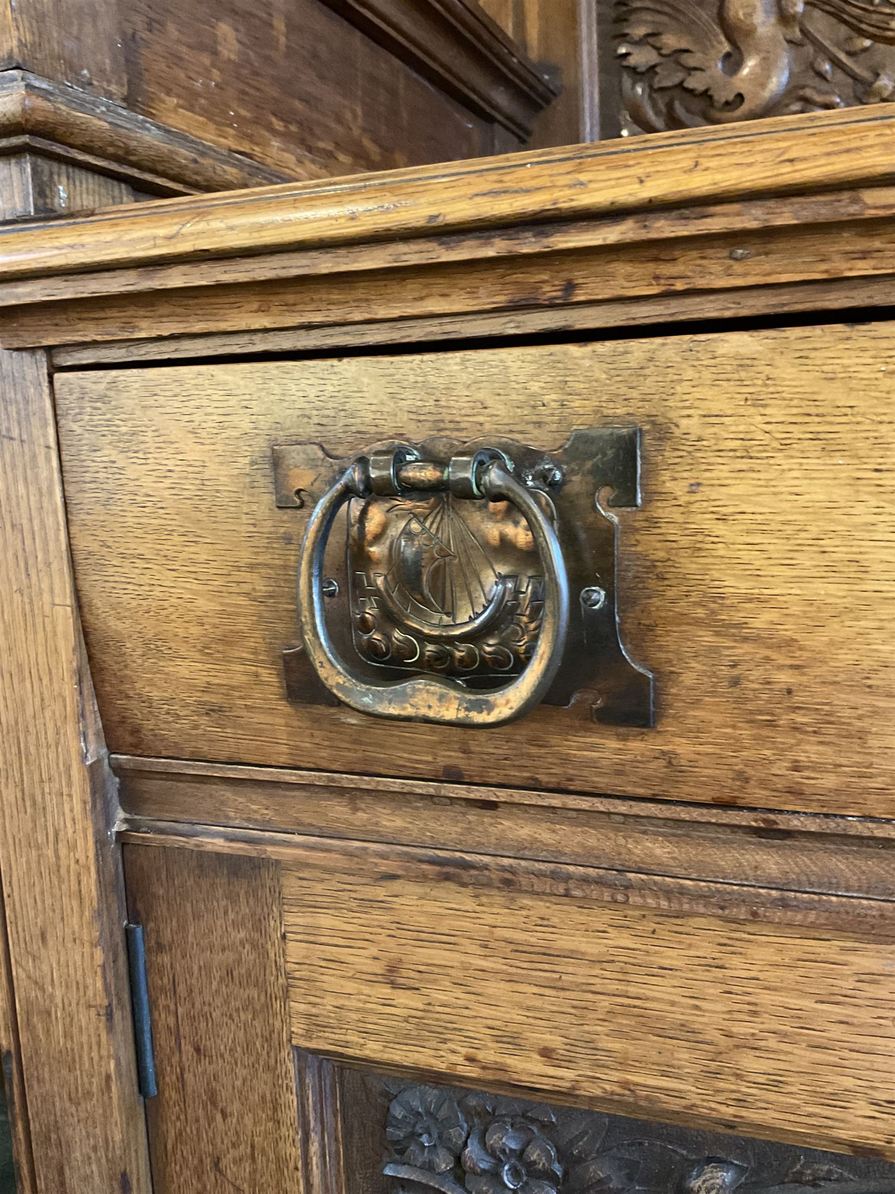 Late 19th century Arts & Crafts period oak mirror back sideboard, shaped bevelled mirror back carved with foliate scrolls and flower heads, down swept supports with elongated club-shaped uprights, moulded rectangular top over two drawers and three cupboards, the drawer fronts with copper handles decorated with sailing ships, panelled doors carved with mythical beasts, flowerheads and foliage, on moulded block feet