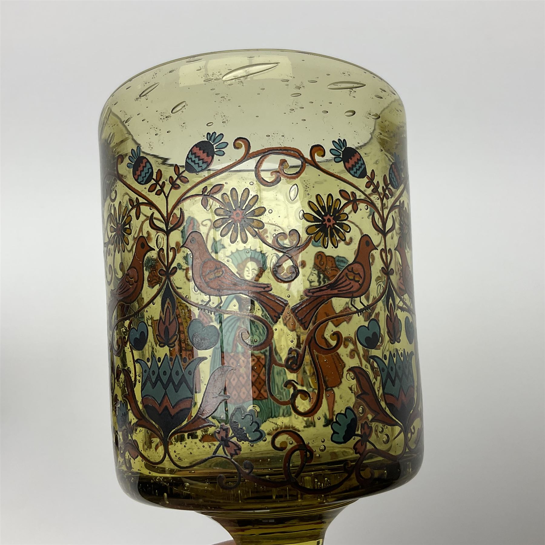 19th Century Bohemian amethyst glass lidded goblet, the bowl of faceted form, gilded with three children in period dress playing games, upon a knopped stem and conforming faceted foot, together with an art nouveau green glass goblet, with gilded rim, upon clear trumpet stem, and a hand blown wine glass, transfer printed with birds and three figures in period dress, tallest H29cm