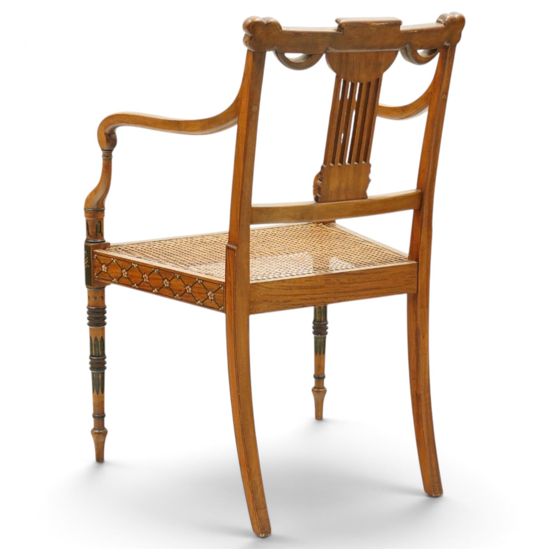 Late 19th century Adam Revival painted and parcel-ebonised satinwood open armchair, the cresting rail centred by a painted panel depicting putti holding garlands, within draped swag ornament and moulded ribbon-twists, over a pierced vertical splat with gilt beaded detail, the downswept arms and caned seat above a painted lattice and floral frieze, raised on ring-turned tapering front supports with musical instrument painted panel and acanthus leaves
