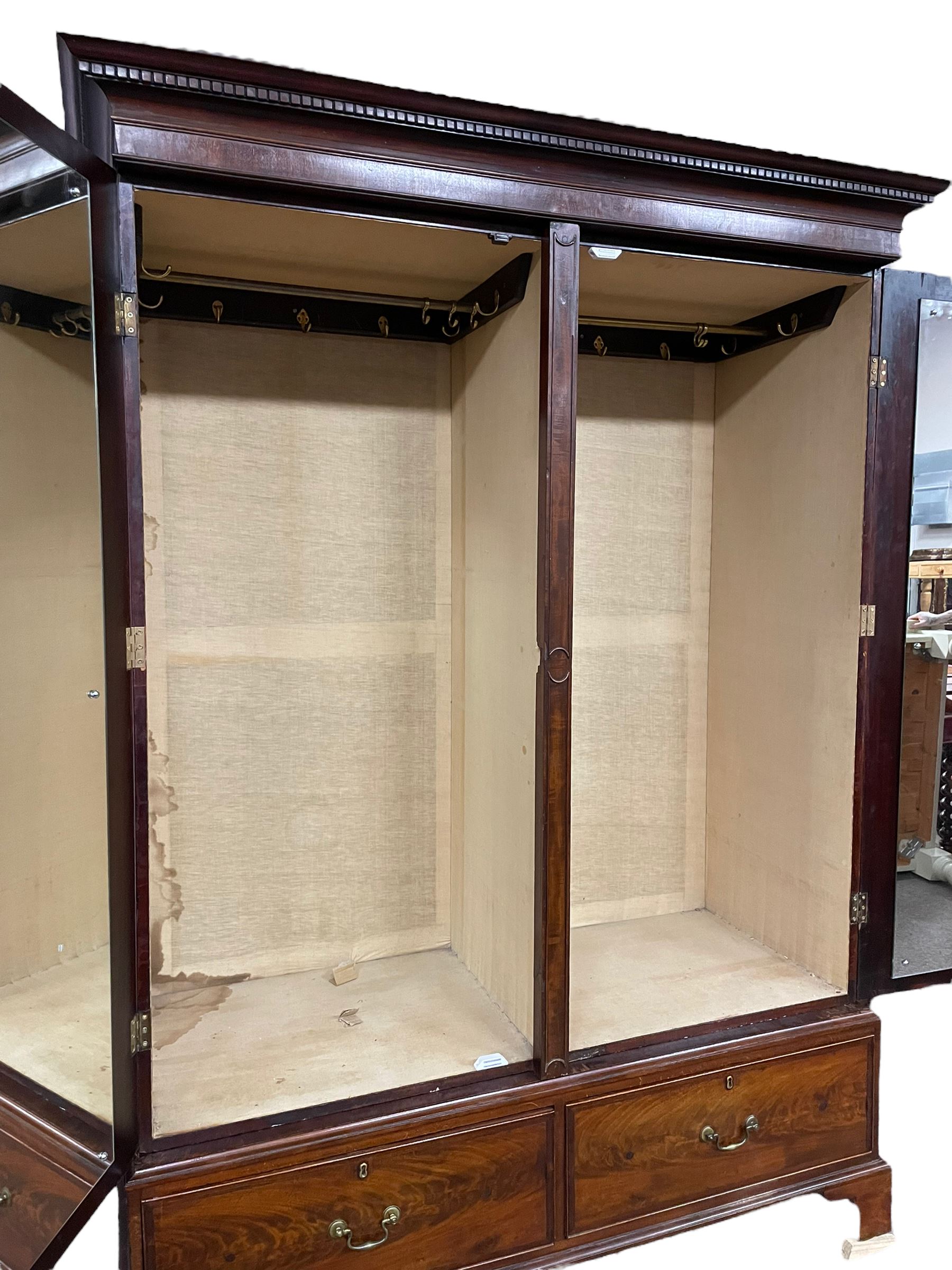 George III mahogany double wardrobe, the projecting cornice with dentil moulding over two panelled doors with mirror-lined interiors, enclosing hanging rails and brass hooks, the base fitted with two drawers, raised on bracket feet