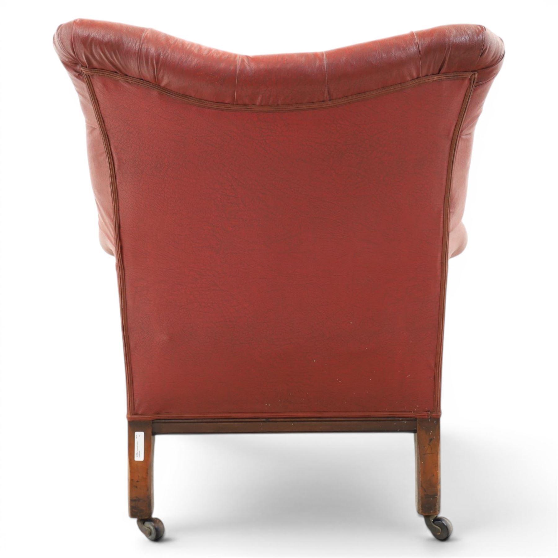 Early 20th century style oxblood red leather Chesterfield armchair, deep buttoned shaped back over padded scroll arms and loose seat cushion, raised on turned mahogany front supports with brass caps and castors
