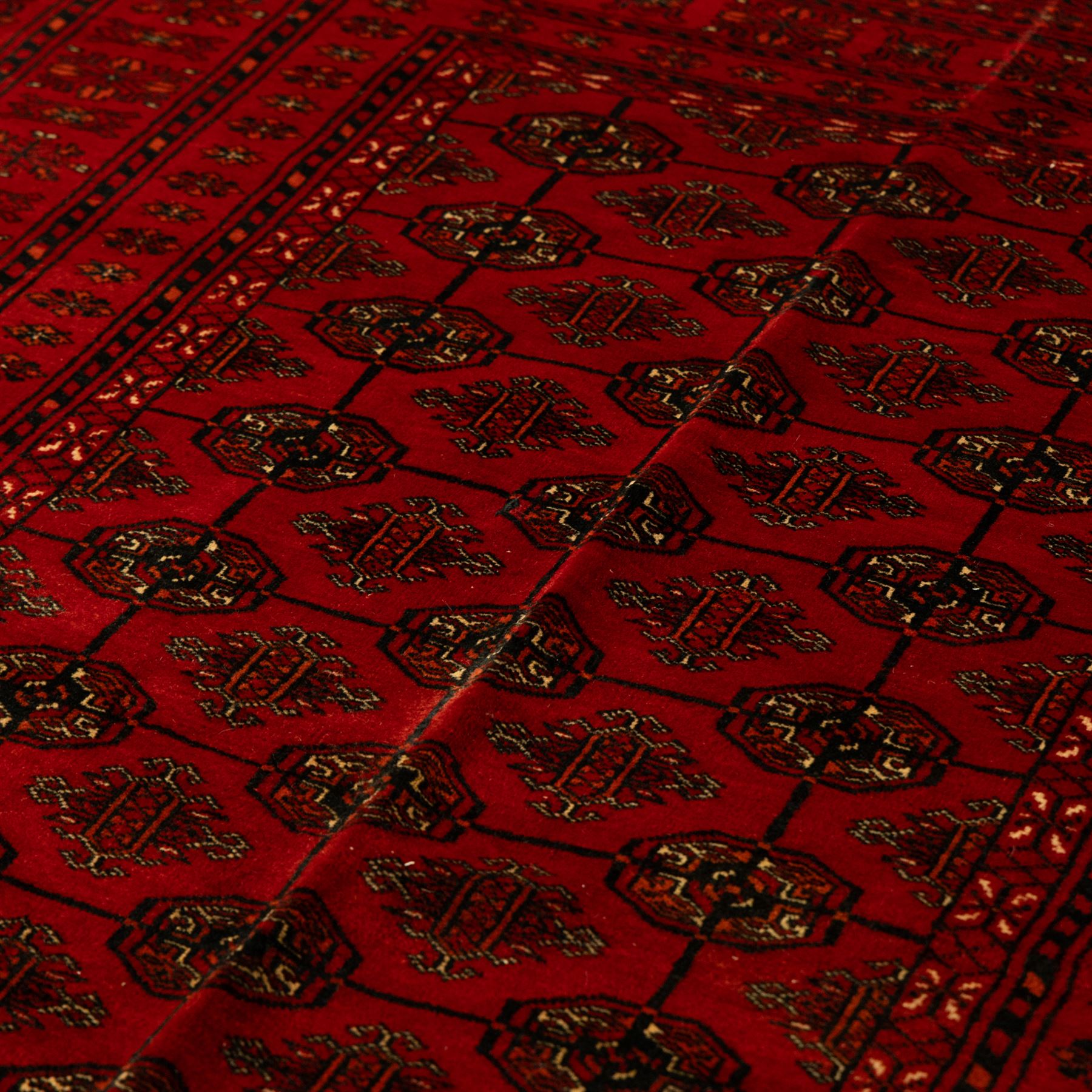 Turkman hand-knotted carpet, deep red ground, with an all-over field of repeating gul motifs arranged in rows, enclosed by multiple red geometric borders with stylised flowerheads and hooked motifs, flanked by narrow patterned guard stripes, with wool fringes