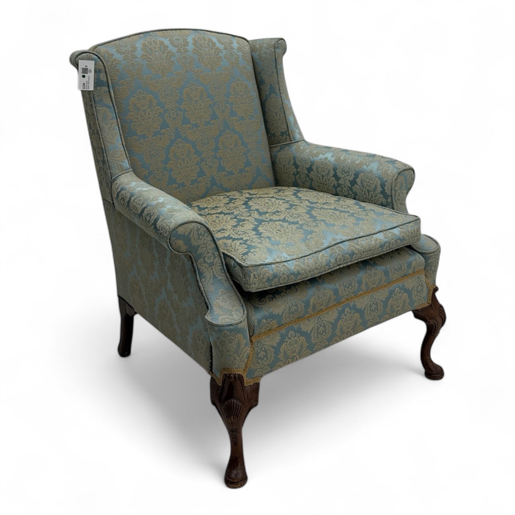 Georgian design carved walnut framed wingback armchair, upholstered in pale blue and gilt floral damask, with shaped loose seat cushion and rolled arms, raised on scallop and cartouche carved front cabriole supports terminating to pad feet