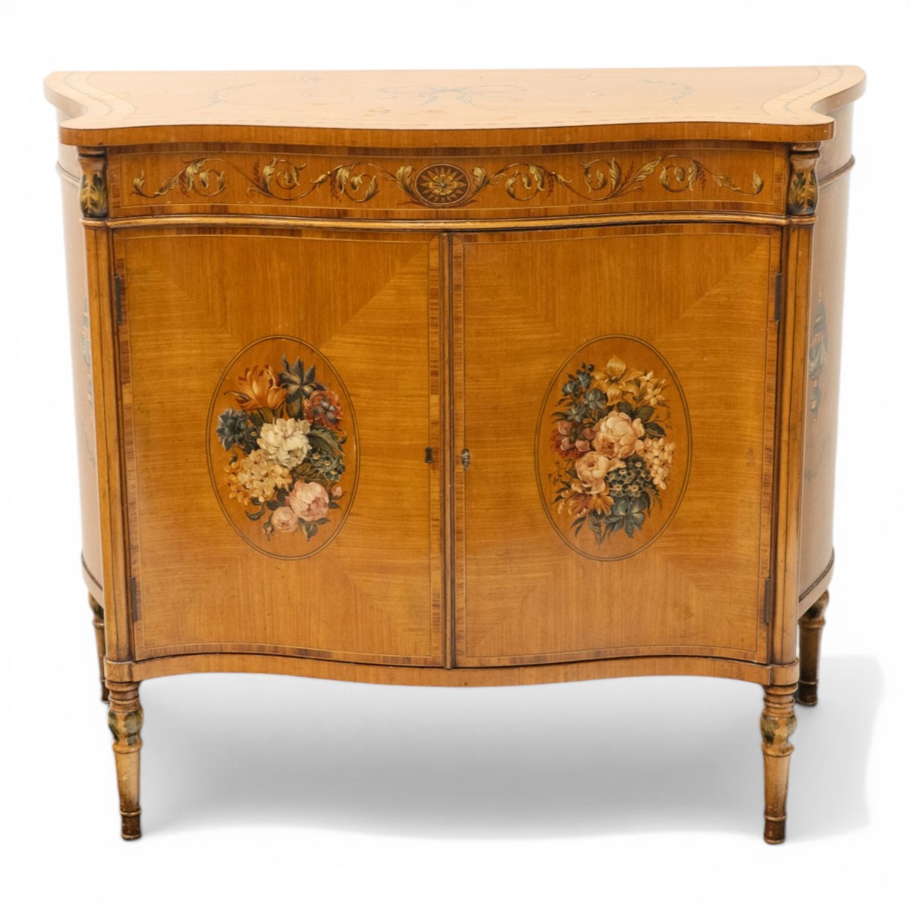 Sheraton Revival satinwood serpentine-front cabinet, the shaped cross-banded top with ebony stringing and painted scrolls of acanthi and ribbon work with a bell-flower border, the banded frieze painted with repeating acanthus scrolls, the matched veneer doors painted with oval panels of floral bouquets, raised on turned and tapering supports