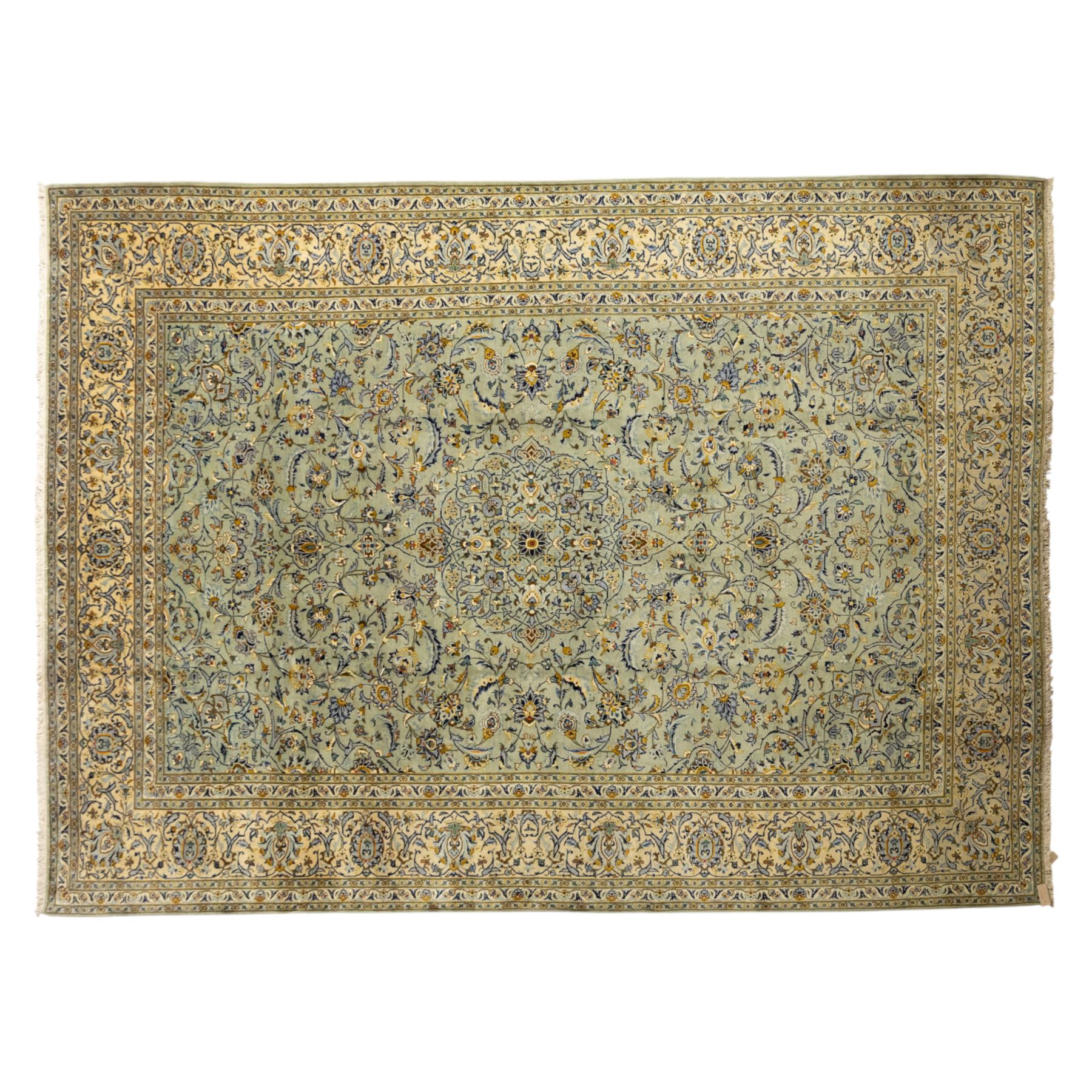 Central Persian Kashan hand-knotted carpet, pale celadon blue ground, decorated with an all-over central floral and foliate design with rosette medallion incorporating scrolling vine work, palmettes, and stylized blossoms  highlighted in tones of indigo, ivory, gold, and powder blue, enclosed by a cream ground border, flanked by multiple guard stripes, with cotton fringe ends 