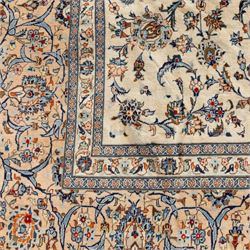Large Central Persian Kashan hand-knotted carpet, ivory field with an all-over scrolling design of vines, palmettes, and floral sprays, enclosed by a broad camel-ground palmette and vine border, framed by multiple guard stripes in a palette of ivory, beige, blue and polychromed tones, with cotton fringe ends 