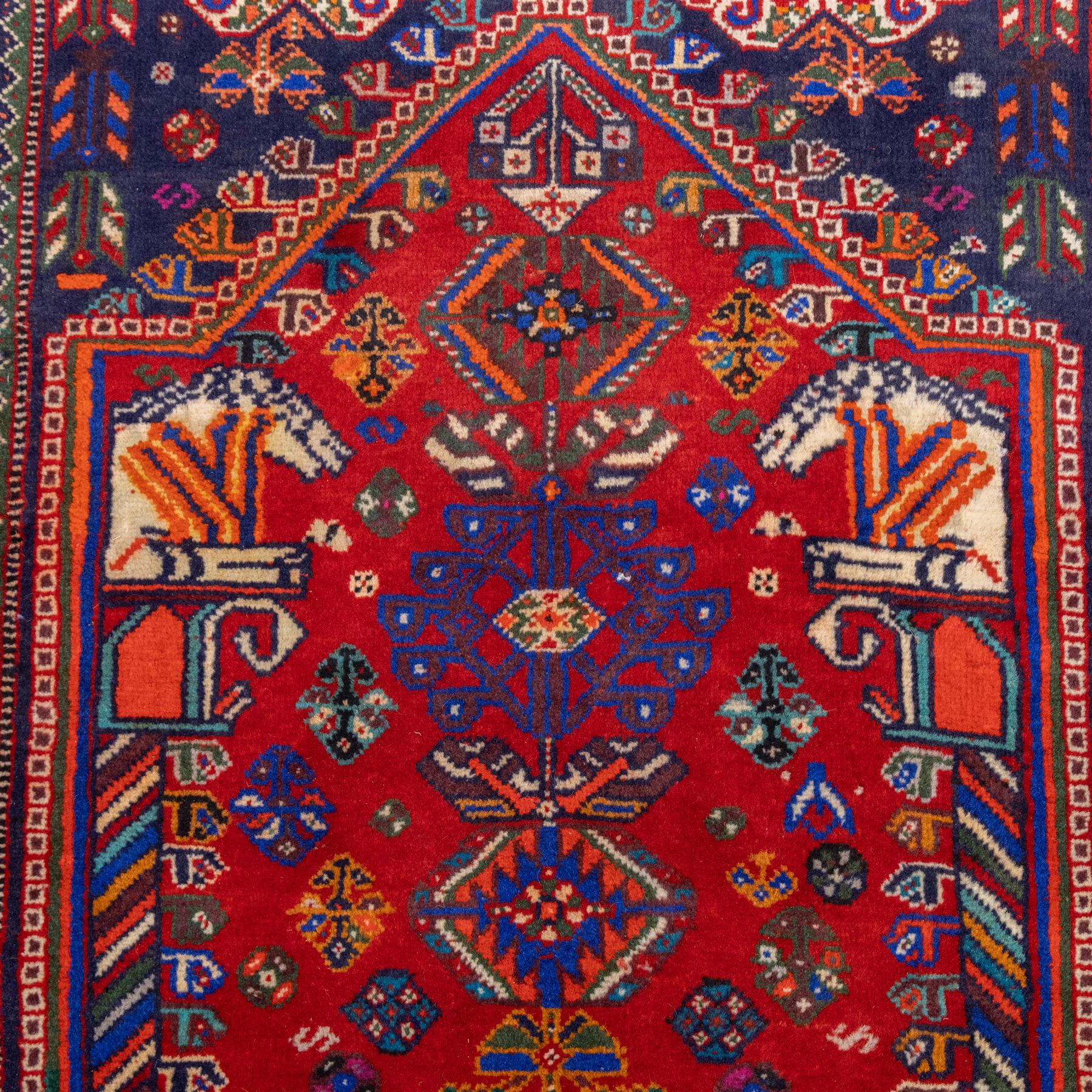 South West Persian Qashqai hand-knotted runner, madder red field  with central vertical succession of stepped lozenge medallions enclosing angular tribal devices and stylized floral motifs, principal medallion in deep indigo with ivory and sky blue highlights, field densely scattered with small geometric ornaments, hooked elements and zoomorphic forms, triangular indigo spandrels enclosed within multiple narrow guard stripes and principal border, with cotton fringe ends 