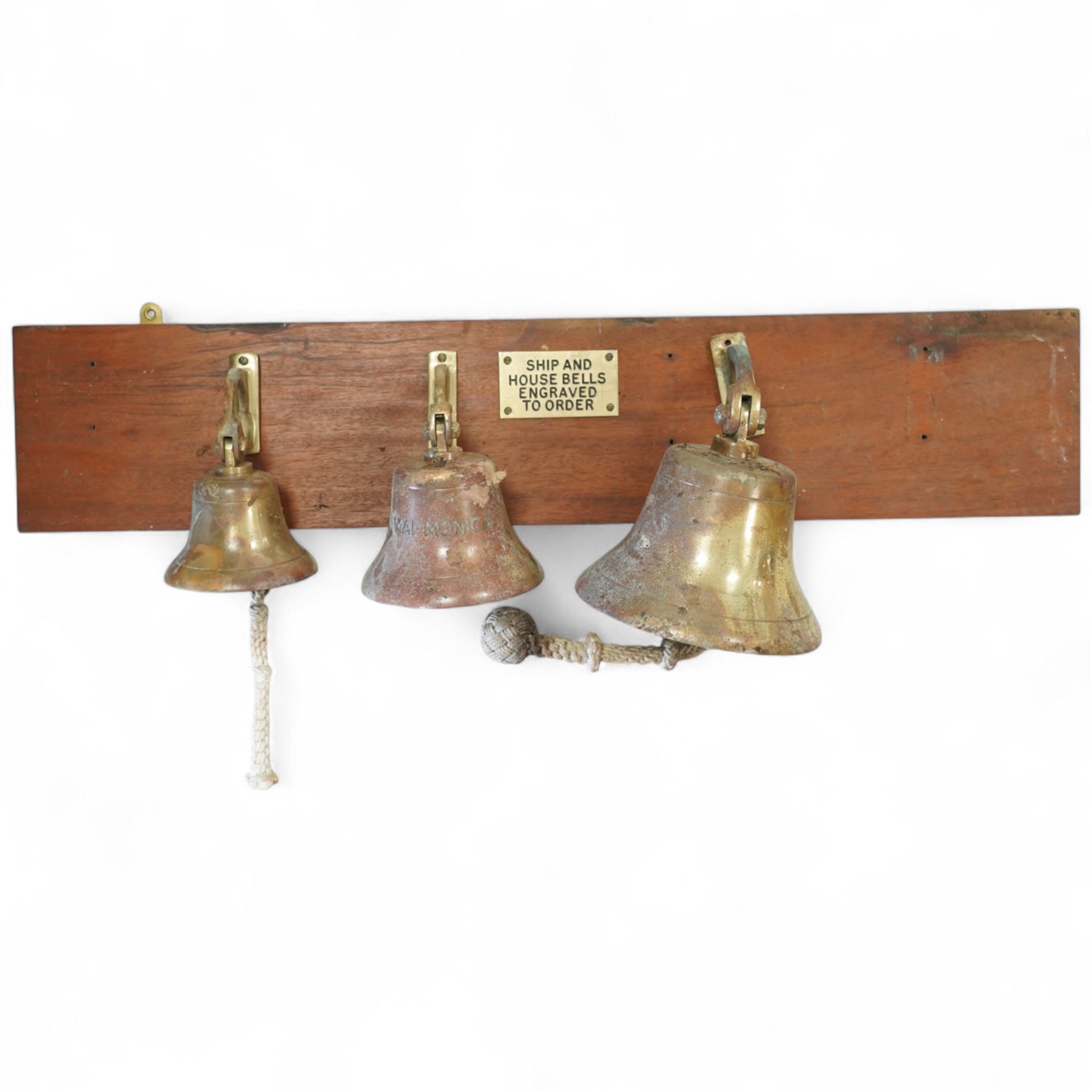 Set of three brass ship bells, each with wall bracket and two with rope pulls, mounted on a rectangular mahogany backboard with brass plaque inscribed ‘Ship and House Bells Engraved to Order’