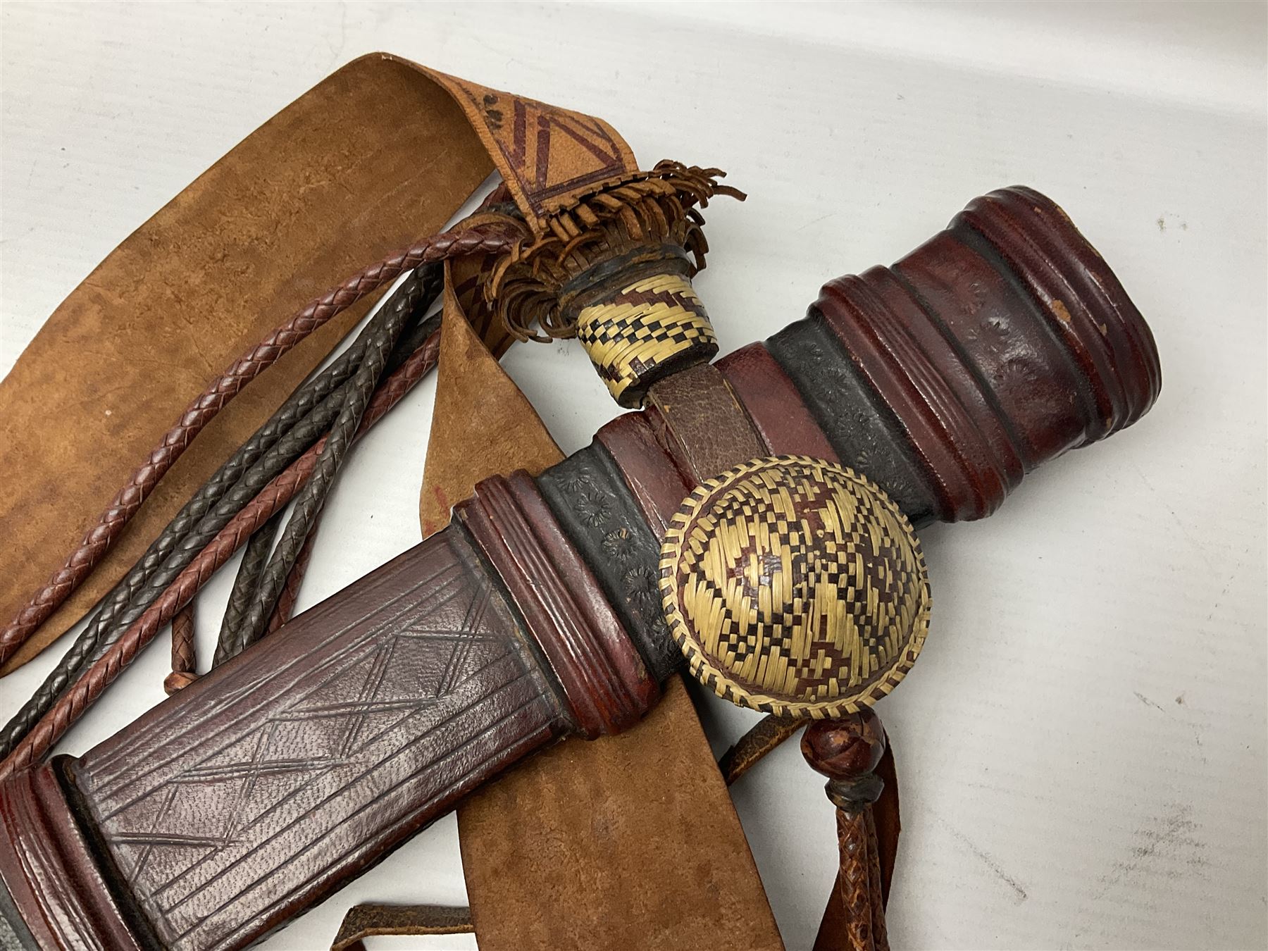 Mandingo sword with 63cm plain curving steel blade and leather covered grip with white metal ribbed spherical pommel; in decorative leather leaf scabbard with plaited tassels and shoulder strap L78cm overall