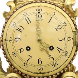 20th century Continental giltwood wall clock, circular dial with Arabic numerals and pierced gilt hands, within a carved case surmounted by a scrolling crest and flanked by stylised dolphins, above a shaped shelf with tassel pendants and a foliate terminal, pendulum missing