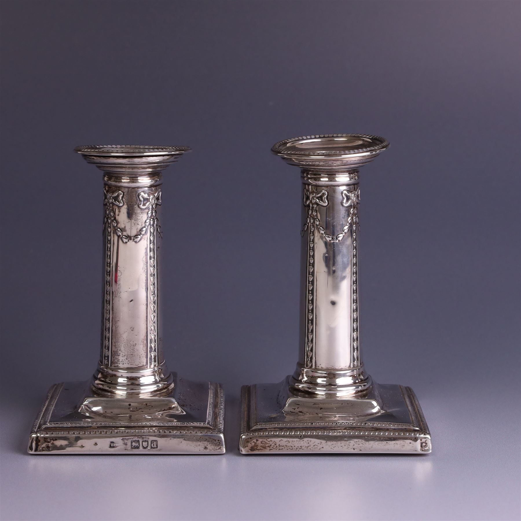 Pair of late Victorian silver mounted candlesticks, the stems embossed with classical ribbon and swag design, upon weighted stepped square bases, hallmarked London 1899, maker's mark worn and indistinct, H12cm