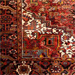 North West Persian Heriz hand-knotted carpet, madder red ground, with a large central geometric medallion with pendants, surrounded by angular foliate and floral motifs incorporating stylised rosettes, palmettes and serrated vine work, enclosed by ivory spandrels and a dark navy border with repeating geometric flowerheads and angular meandering vines, flanked by narrow patterned guard stripes, with cotton fringes