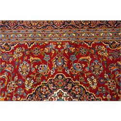 Central Persian Kashan hand-knotted carpet, crimson field with central cusped medallion in ivory, pale blue and indigo, issuing a network of scrolling arabesques, palmettes, and floral sprays, with conforming spandrels, main border of scrolling vinery and stylized palmettes, framed by multiple guard borders, with cotton fringe ends 