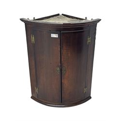 Early 19th century oak bow fronted wall hanging corner cupboard, projecting cornice over double doors enclosing shelving, on plinth base 