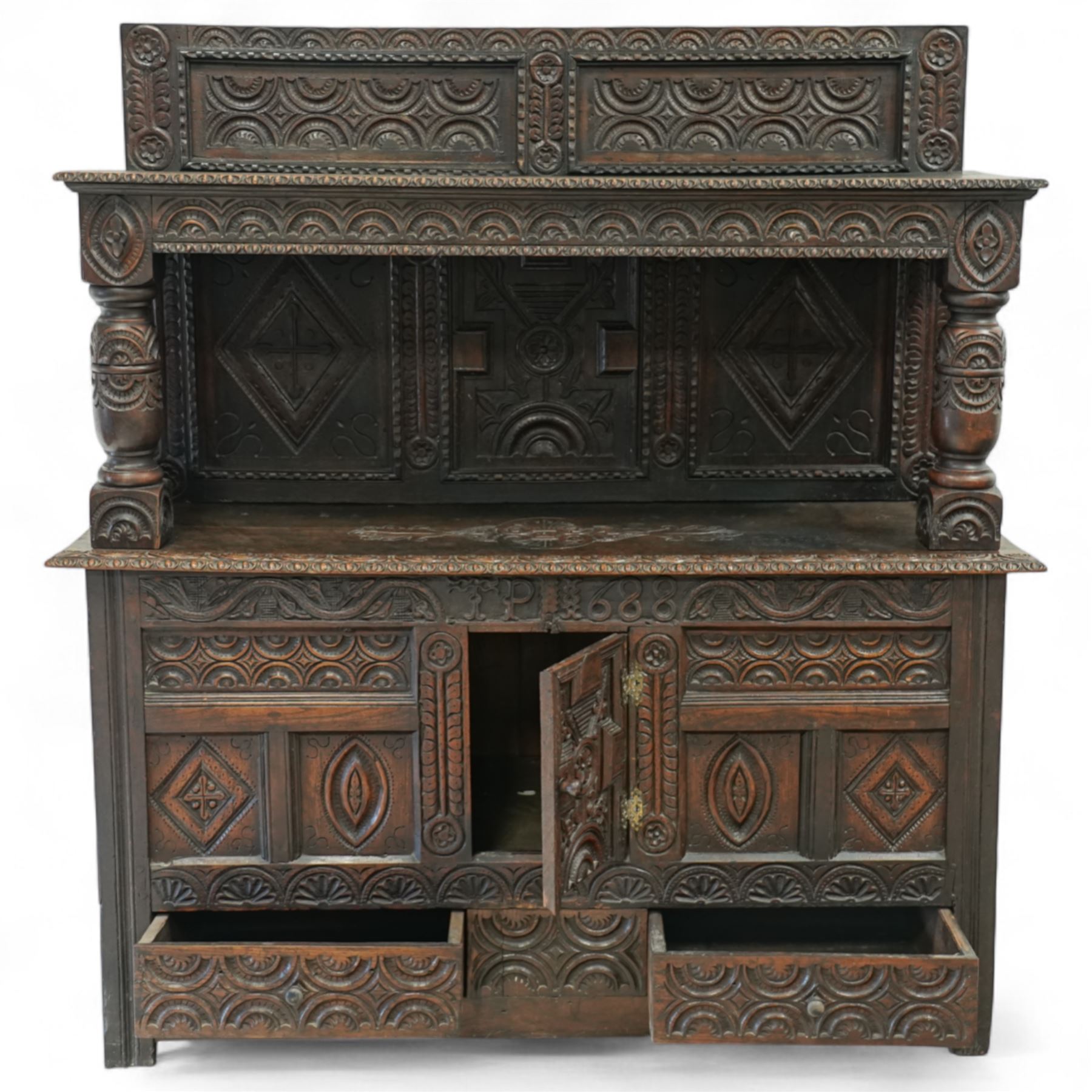 An early 20th-century Jacobean revival style carved oak Court Cupboard, tier-form of 17th-century design, rectangular form with carved back with geometric motifs and arches, projecting top section over twin baluster columns connecting the lower tier above a carved panelled front and panelled cupboard door with profusely carved lozenges and lunettes, above two base drawers, raised on block feet 