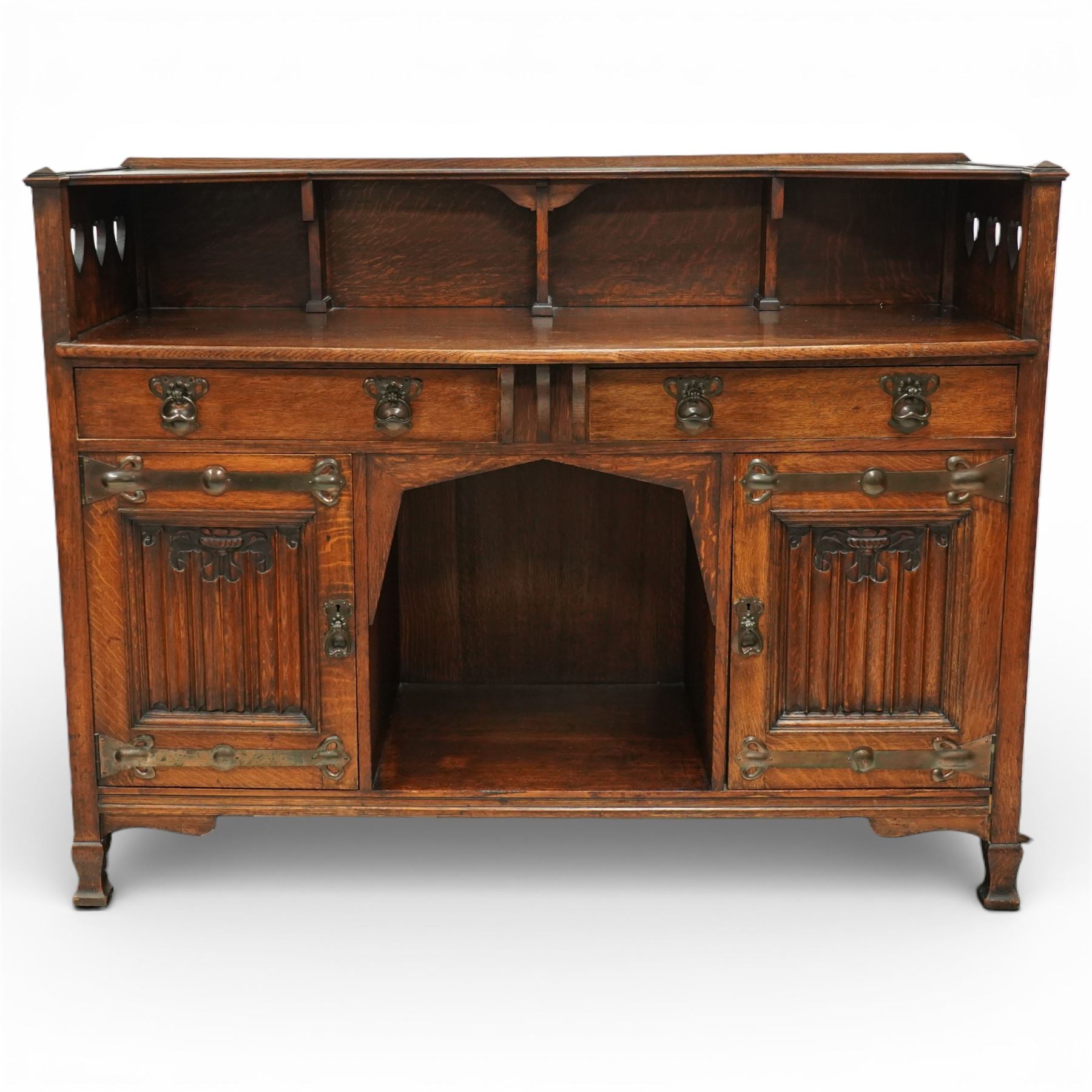 Shapland and Petter - Arts and crafts oak sideboard, rectangular overhanging top with raised open back fitted with shaped supports and pierced heart cut-outs to the ends, the base fitted with a central arrangement of two short drawers with cast metal drop handles over an open kneehole recess, flanked by a pair of cupboard doors each with linenfold panels and applied strap hinges, enclosing shelved interiors, raised on shaped bracket feet