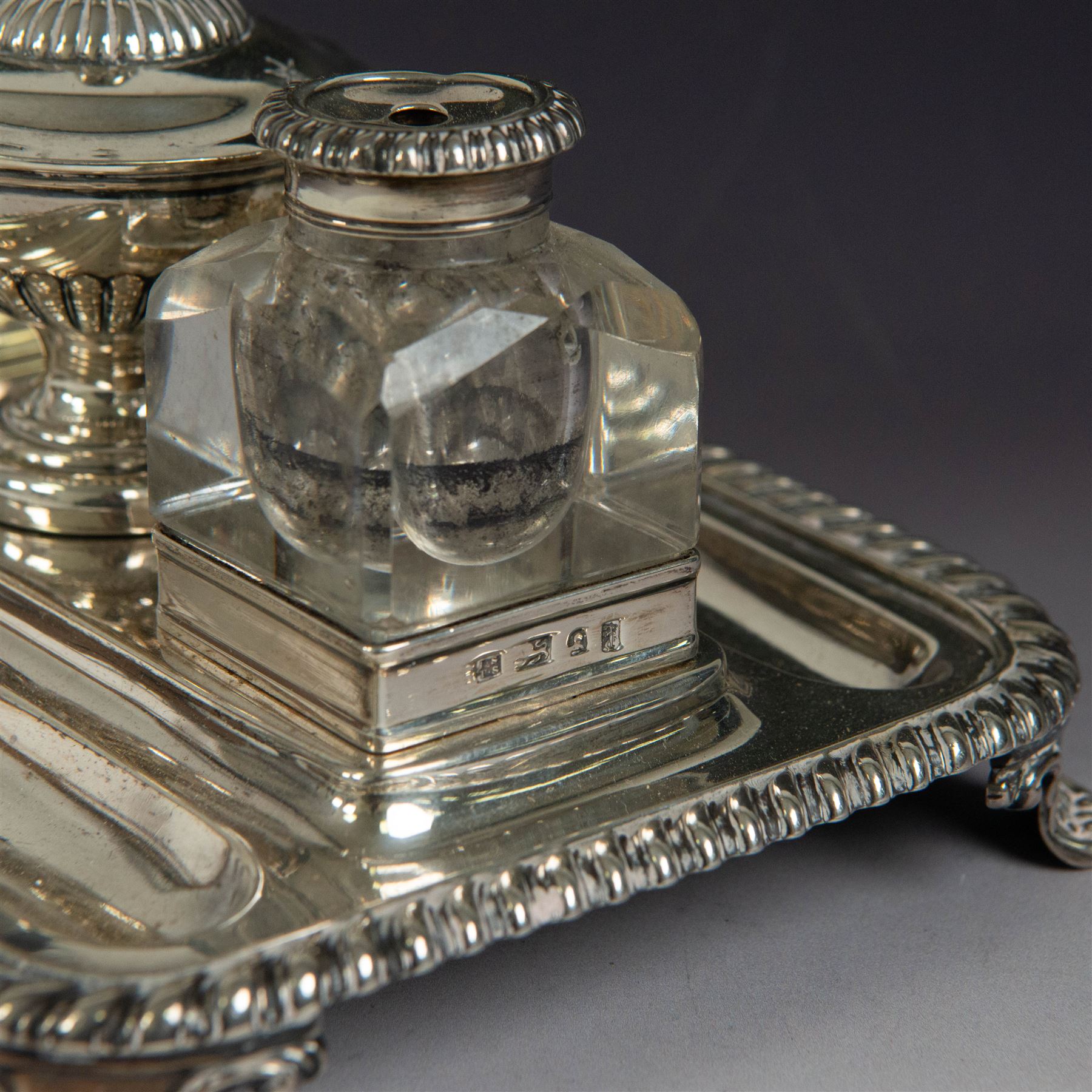 George IV silver desk stand, of rectangular form with rounded corners and oblique gadrooned rim, upon four embossed pad feet, with central pedestal dish of part fluted form with domed lid engraved with crest and with bud finial, with two recesses either side housing faceted glass inkwells, each with silver collars, hallmarked John & Thomas Settle, Sheffield 1825 & 1827, H11cm
