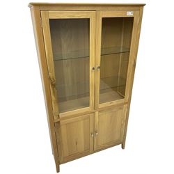 Contemporary light oak display cabinet on cupboard, fitted with two glazed doors enclosing two adjustable glass shelves with light fittings to the top, over panelled cupboard concealing single shelf, on square supports