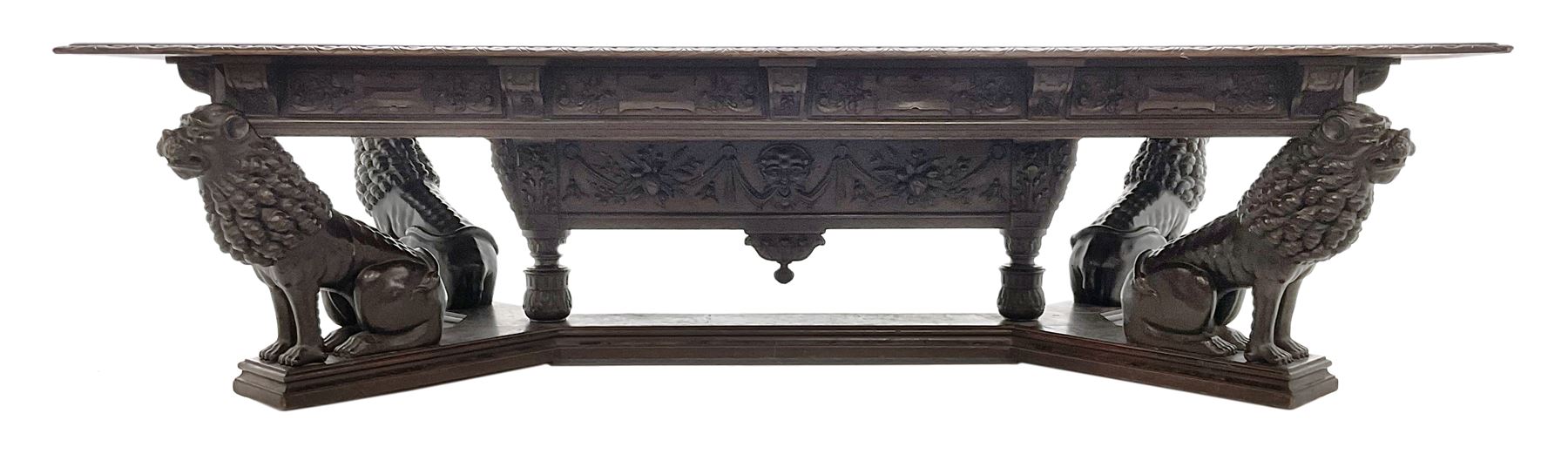 Large 19th century carved oak library table, the rectangular top with lunette carved edge over cartouche panelled and carved  scroll frieze, the central tapering rectangular support relief carved with masks and garlands, on four carved seated lion supports at each corner, on flat angular stretcher with recessed castors stamped ‘Bartholomew & Fletcher, London’