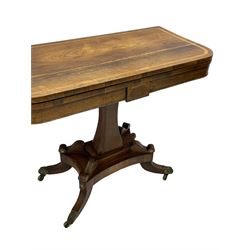Regency rosewood and brass inlaid card table, the rectangular fold-over and swivelling top with rounded corners with a satinwood band, opening to reveal a blue baize lined interior playing surface, the frieze with a raised panel with satinwood stringing and a lower beaded brass edge, the concave and canted column with a shaped platform base applied with scrolls with brass star inlays and stringing, quadruple splayed supports terminating in foliate cast cups and castors