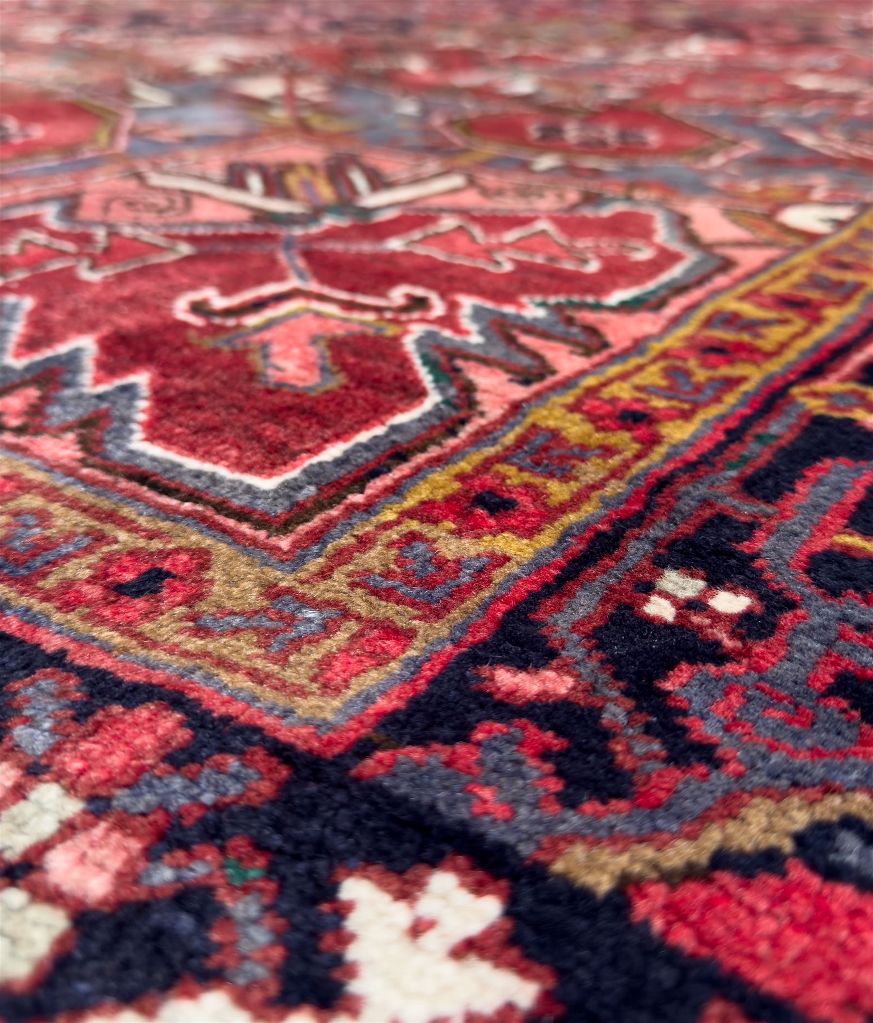 Northwestern Persian Heriz hand-knotted carpet, madder red field with a bold stepped medallion in indigo, ivory, and ivory, surrounded by angular floral motifs and stylized leaves, and the spandrels set within a dark border of rosettes and angular vine motifs, 335cmx252cm
