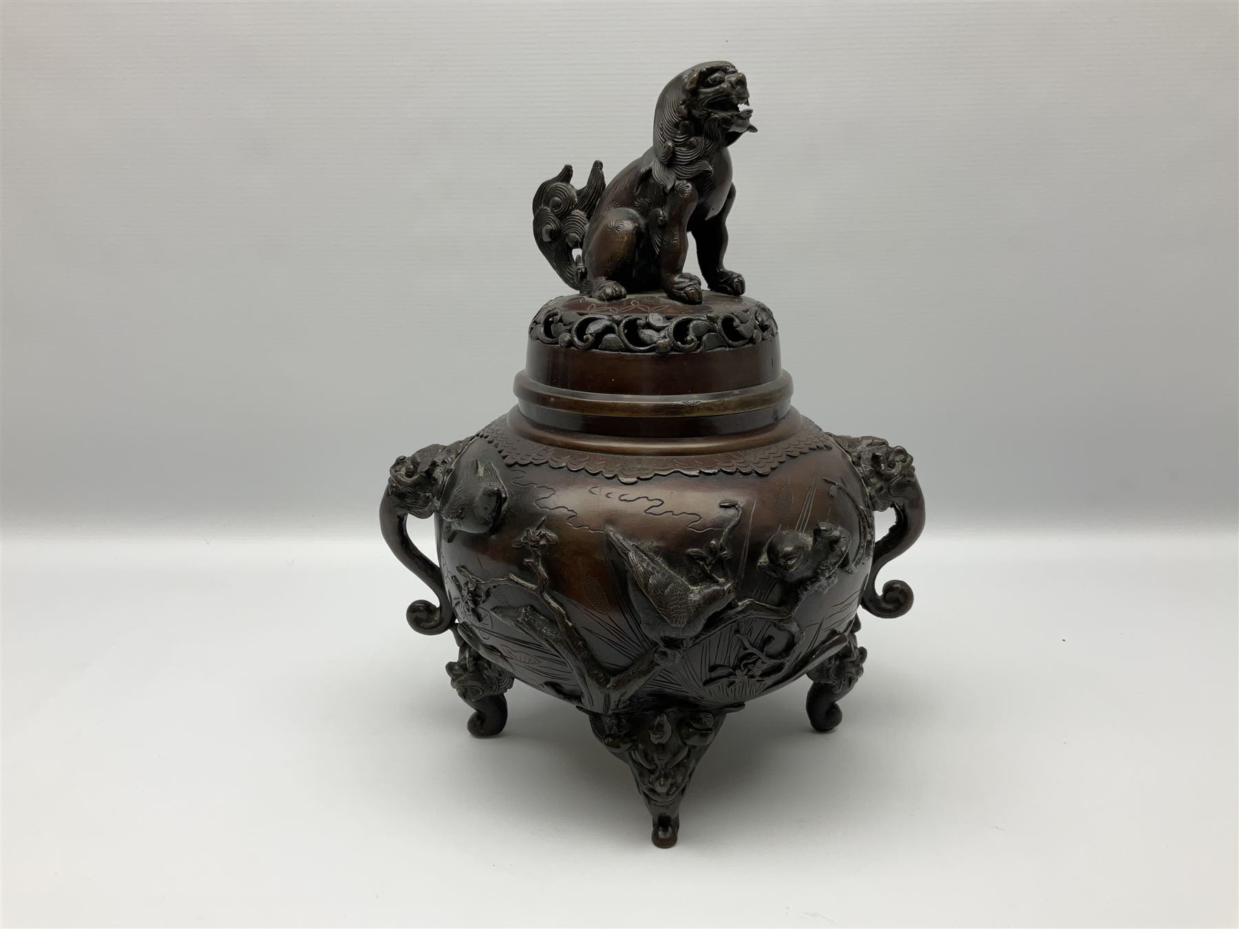 Japanese bronzed koro, the body of globular form with twin cast zoomorphic mask handles, decorated in relief with birds upon blossoming branches, against a ground incised with rushes and peonies, the flat toped domed cover with pierced band and surmounted by a cast figure of a Foo dog, the whole upon three zoomorphic mask feet, with impressed character type mark beneath, H35.5cm