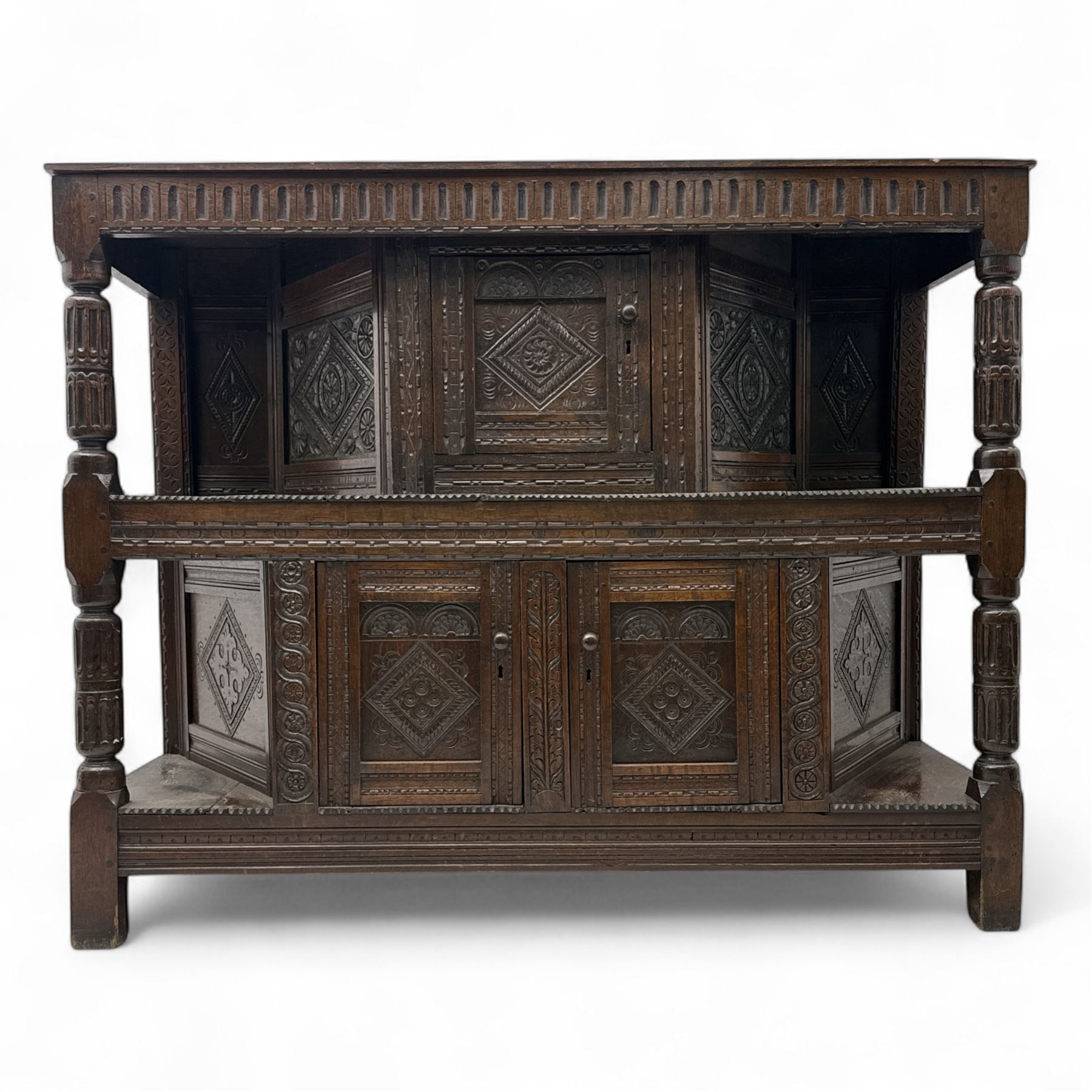 Early 20th-century  oak Jacobean two-tier revival court cupboard, rectangular top with carved notch and stylized foliate carved edge above a carved lozenge pattern frieze and a central cupboard with carved geometric designs and geometric carved panelled sides, above a carved panelled cupboard with two doors of similar geometric design, the whole flanked by two sets of stop-fluted baluster turned columns and supported on stile feet