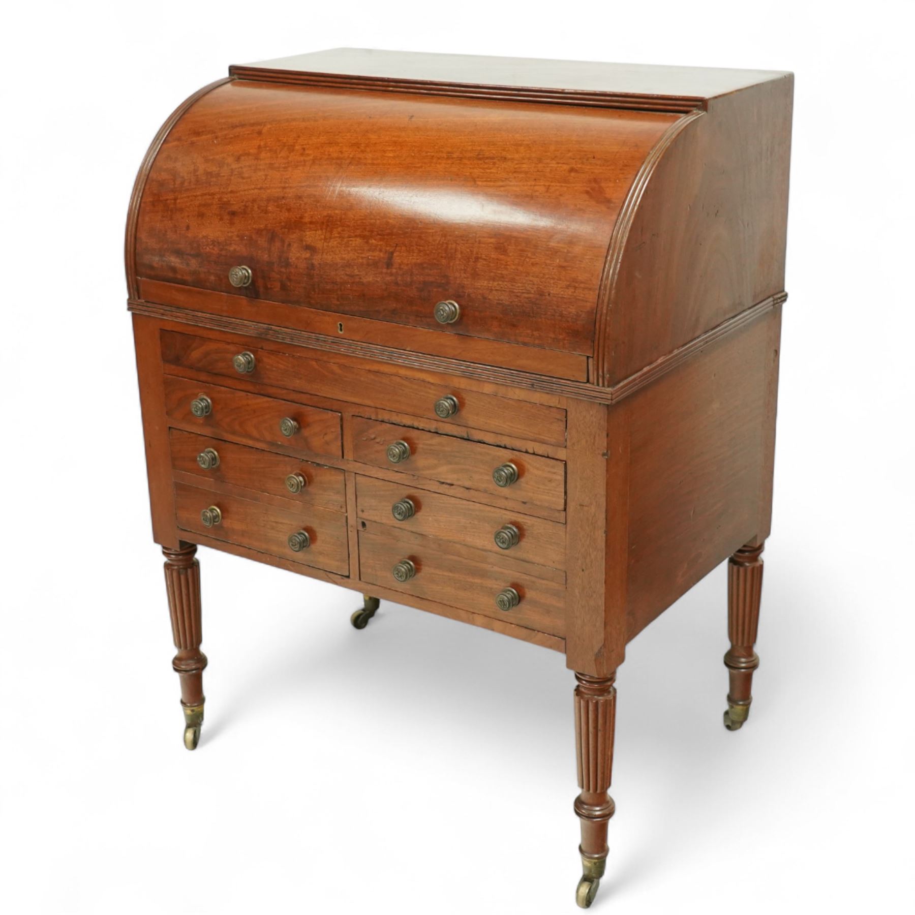 Regency period mahogany cylinder desk, the roll-top enclosing a fitted satinwood interior with pigeonholes and drawers, sliding green baize writing surface, lower section fitted with four drawers and a cupboard, on reeded tapering legs with brass caps and castors