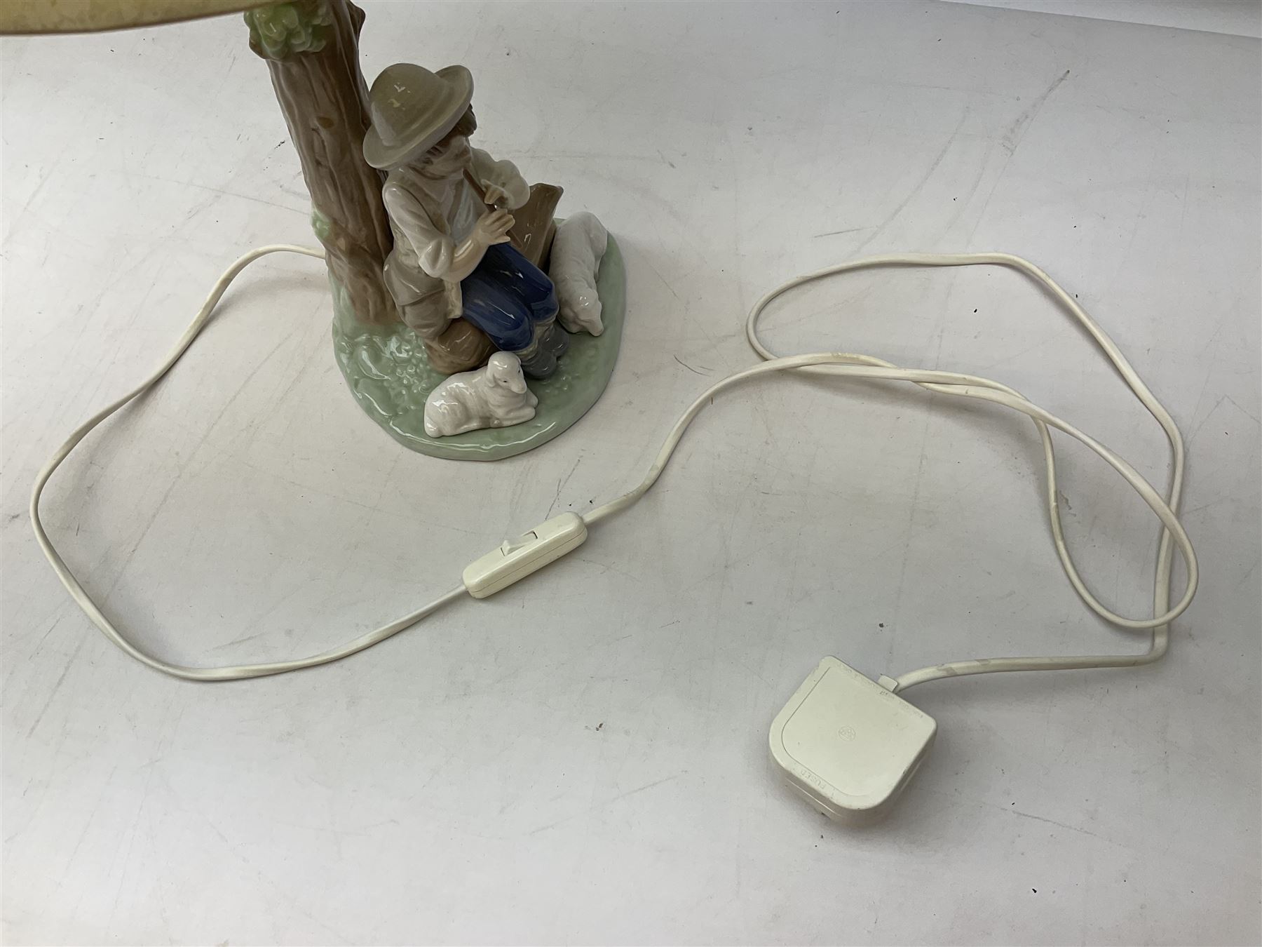 Nao table lamp modelled as a boy playing the flute upon logs with two recumbent sheep at his feet, the stem modelled as a tree trunk, with fabric shade, with printed mark beneath, H46cm