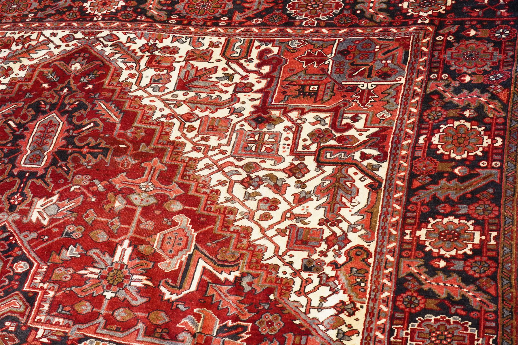 North West Persian Heriz hand-knotted carpet, brick red field with a large central angular star medallion with pendants surrounded by stylized geometric palmettes, serrated leaves and tribal motifs,, ivory corner spandrels, field enclosed by a dark indigo main border of stylize rosettes and angular vine motifs between multiple guard stripes, with cotton fringe ends 