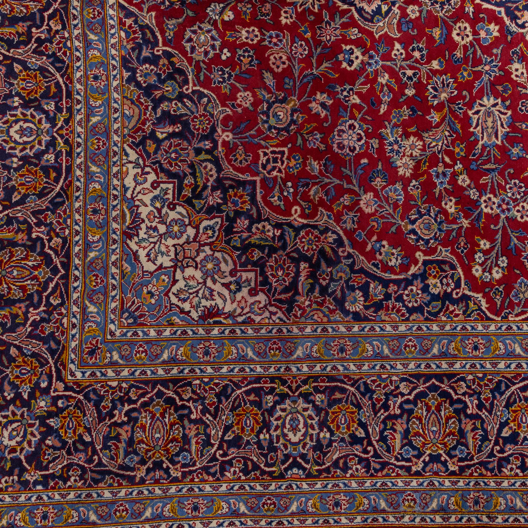 Central Persian Kashan hand-knotted carpet, madder red field with central cusped medallion in navy and ivory enclosing a floral rosette and radiating scrolling arabesques and palmettes, across a densely ornamented ground of finely drawn vinery enriched with a profusion of flowering sprays and stylized blossoms, spandrels in deep indigo and principal border of navy ground decorated with a continuous palmette and vine-scroll flanked by multiple guard stripes, with cotton fringe ends