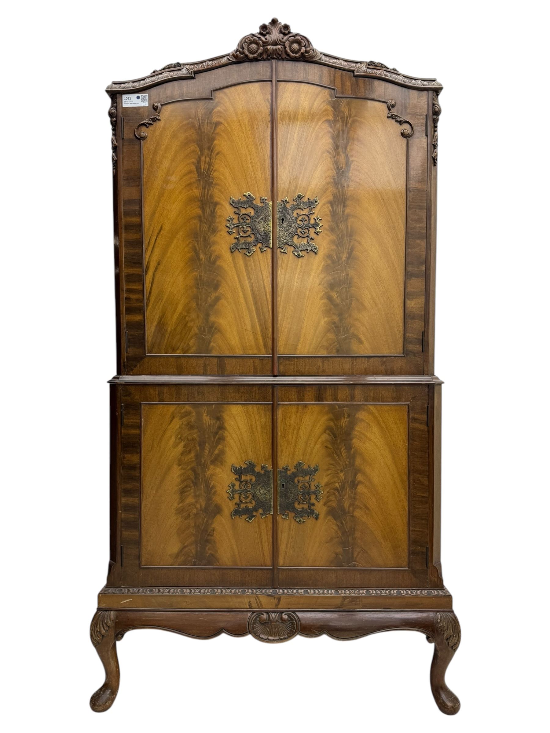 Early 20th century walnut veneered cocktail cabinet, shaped and carved foliate pediment above a pair of arched panelled doors with figured veneers and ornate cast escutcheons, interior is mirrored on the base and back, slide out mirrored shelf, flanked by foliate carved uprights, over a conforming lower cabinet, all raised on shaped apron and scroll carved cabriole feet