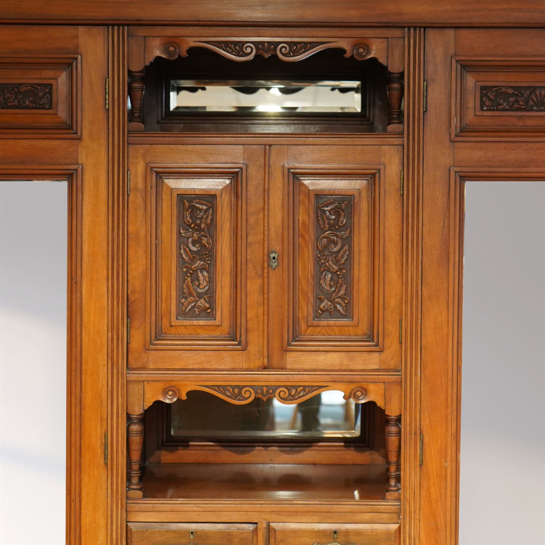 Edwardian walnut triple combination wardrobe, projecting pediment with pierced foliate scroll cresting above a central cupboard flanked by bevelled mirrored doors, the central section with twin panelled cupboard doors carved with foliate scrolls, over open recess with mirrored back and turned supports, above two short and three long drawers with brass handles, interior fitted with hanging rails and shelf, raised on plinth base