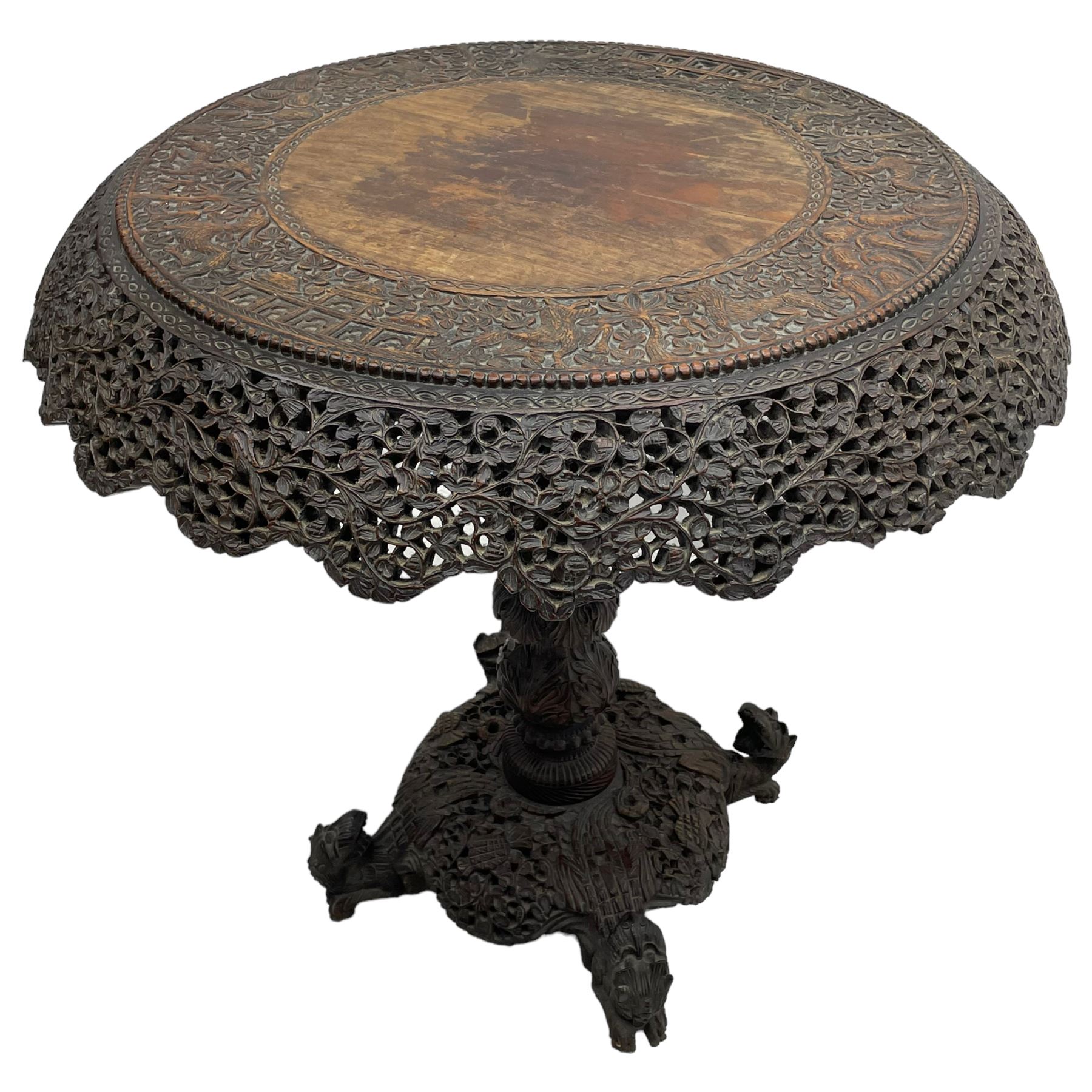 Late 19th century Anglo-Indian carved hardwood occasional table, circular tilt-top carved profusely with mythical animals and foliage decoration, pierced and carved with interlaced leafy branches, leaf carved pedestal supports on a raised platform with further carved and pierced foliage decoration, four carved feet in the form of mythical beasts with gaping jaws 
