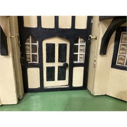 Large Tri-ang No.93 doll`s house in the 'Stockbroker's Range' c1935; two-storey double-fronted Tudor design, half timbered gables, two-storey bay windows, cream and mock shrubbery to front, opening metal framed windows with green shutters, red simulated tiled roof with chimney, integral garage with opening doors, front elevation has four hinged doors opening to reveal two bedrooms with fire places, living room, bathroom and kitchen with built in dresser and sink, entrance hall and staircase, side entrance with porch and seat and sun dial over on chimney stack; some wiring for electrical lighting; metal Tri-ang Toys disc verso; comprehensively furnished H66cm L117cm D43cm