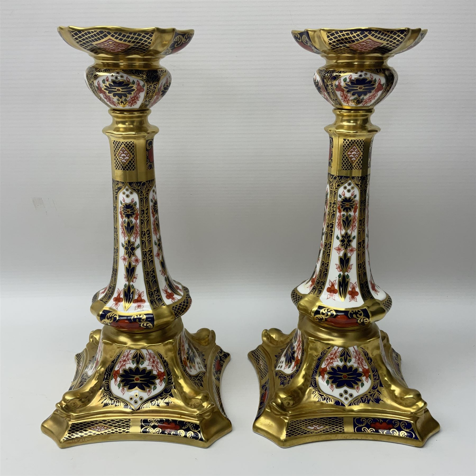 Pair of modern Royal Crown Derby Old Imari 1128 pattern candlesticks, each with shaped drip tray affixed to squat socket, upon spreading stem and canted square base with gilt dolphin edges, with printed marks beneath including Roman numeral year cypher for 2003, H26.5cm, with original boxes