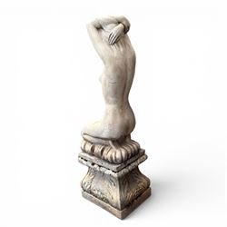 20th century composite stone garden figure, modelled as a kneeling nude female with arms raised behind her head, mounted on an integral cushion, the squared ogee pedestal moulded with acanthus leaves and flower heads, in a white painted and weathered finish