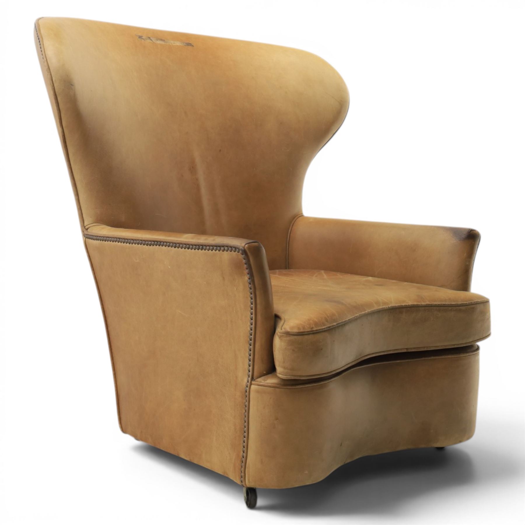 20th century tan leather armchair, the curved wing back above low square arms, shaped loose seat cushion with piped edge, close-nailed side detail, raised on concealed castors