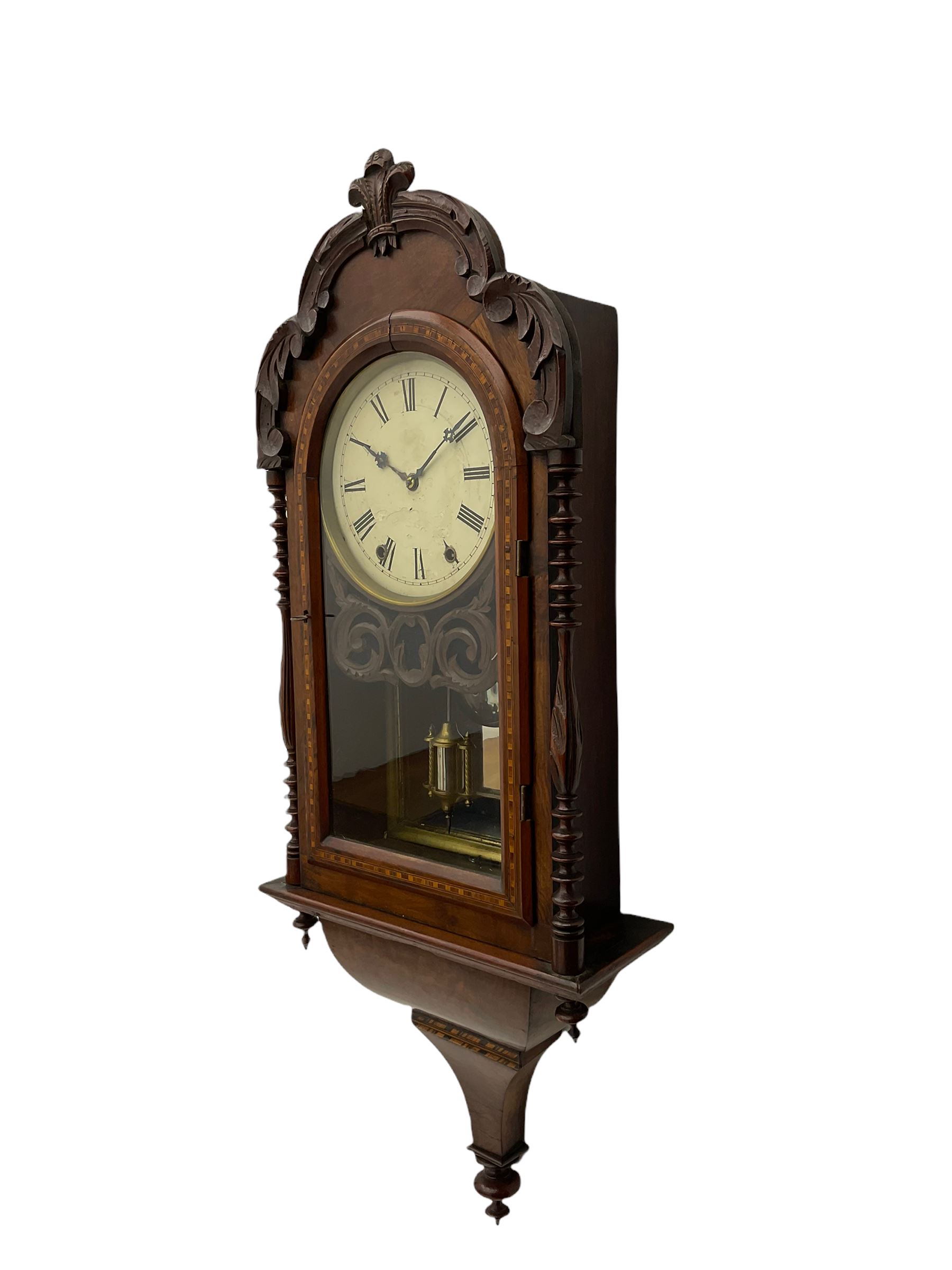 A 19thcentury American wall clock in a walnut case with crossbanding, with a carved pediment surround and arched glazed door flanked by two ring turned columns with pendants on an ogee plinth beneath, with a spring driven movement striking the hours on a bell, eight-inch painted dial with roman numerals, minute track and steel Maltese-Cross hands, with a spun brass bezel and winding collets, visible mock mercury pendulum with a reflective silvered mirror to the rear. With key and pendulum.   

