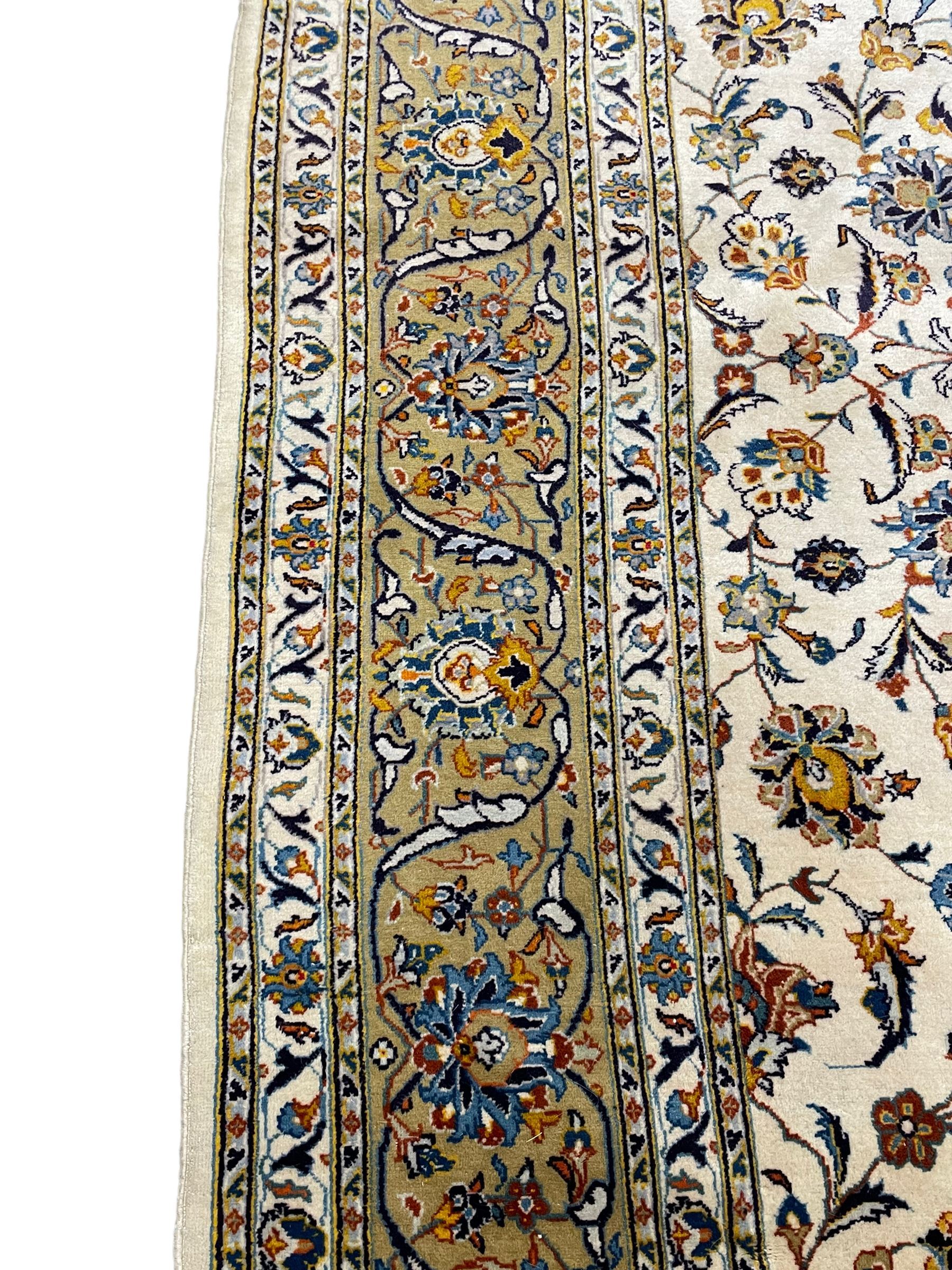 Persian Kashan ivory ground carpet, the field decorated with trailing foliate branches and stylised flower head motifs, guarded border with repeating design decorated with scrolling leafy branch and plant motifs 