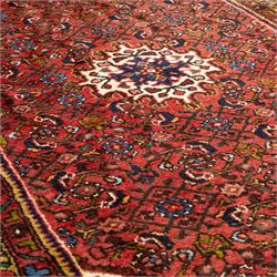 Hamadan hand-knotted rug, red field with small central floral medallion and dense repeating geometric motifs in dark blue, cream and green tones, within multiple patterned borders