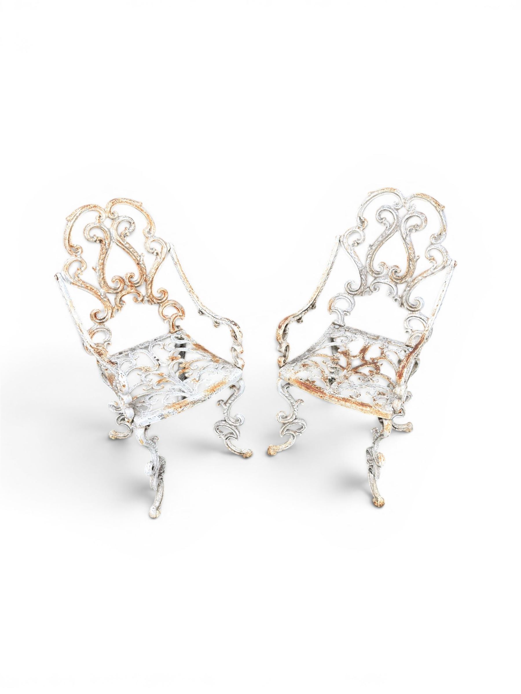 Pair of Victorian cast iron garden chairs, the ornate pierced back of scrolling foliate design, scrolling arm terminals over splayed supports formed of C and S-scrolls, in distressed white painted finish