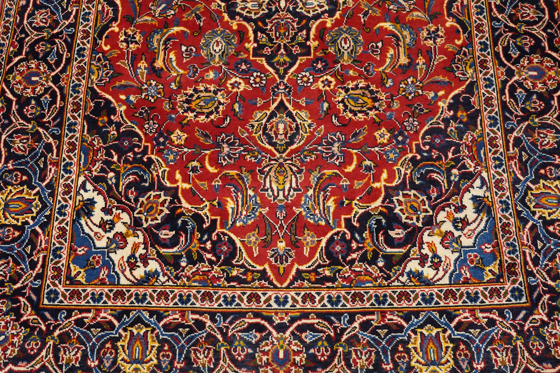 Central Persian Kashan hand-knotted rug, madder red field, with central lobed medallion with pendants, issuing an all-over design of scrolling vine, palmettes, and floral sprays, with conforming spandrels, enclosed by a broad indigo palmette and vine border framed by guard stripes in a palette of navy, blue and ivory tones, with cotton fringe ends 